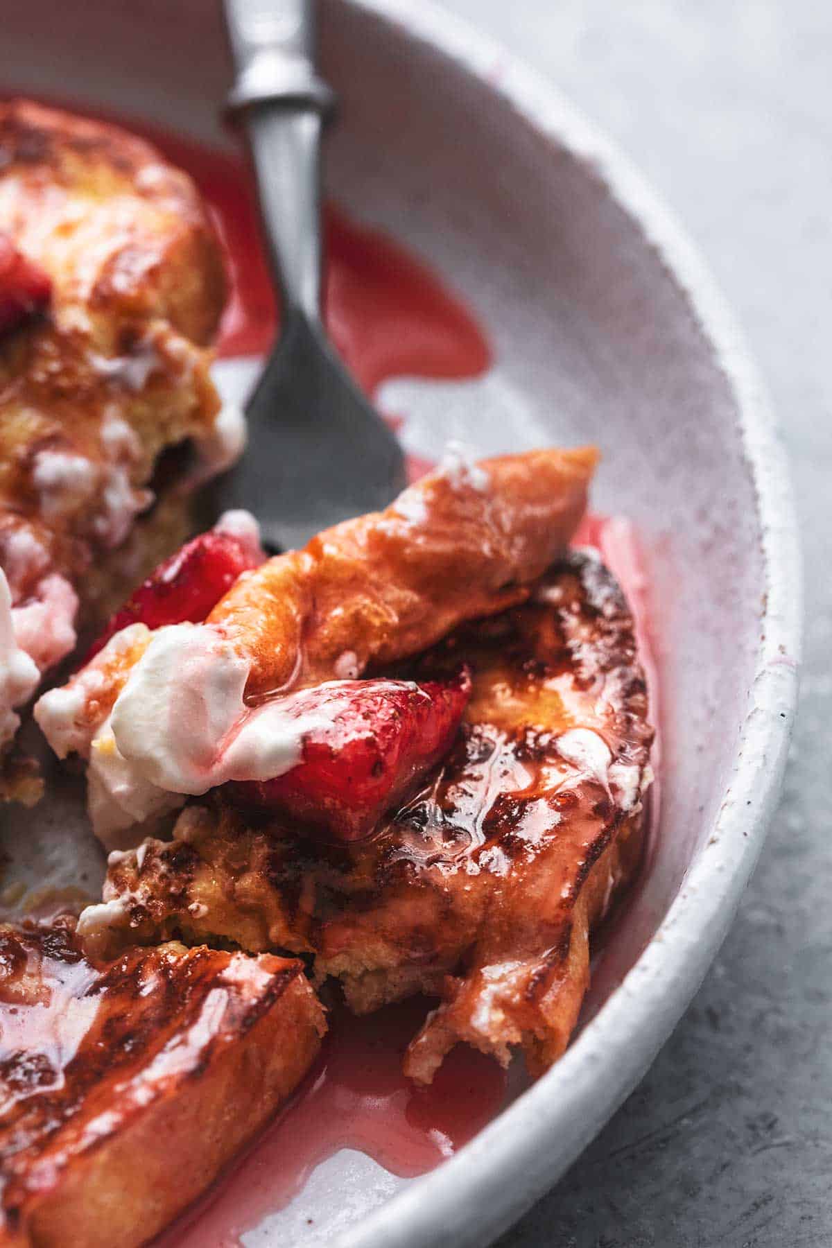 close up of a bite of strawberries and cream French toast on a fork on a plate.
