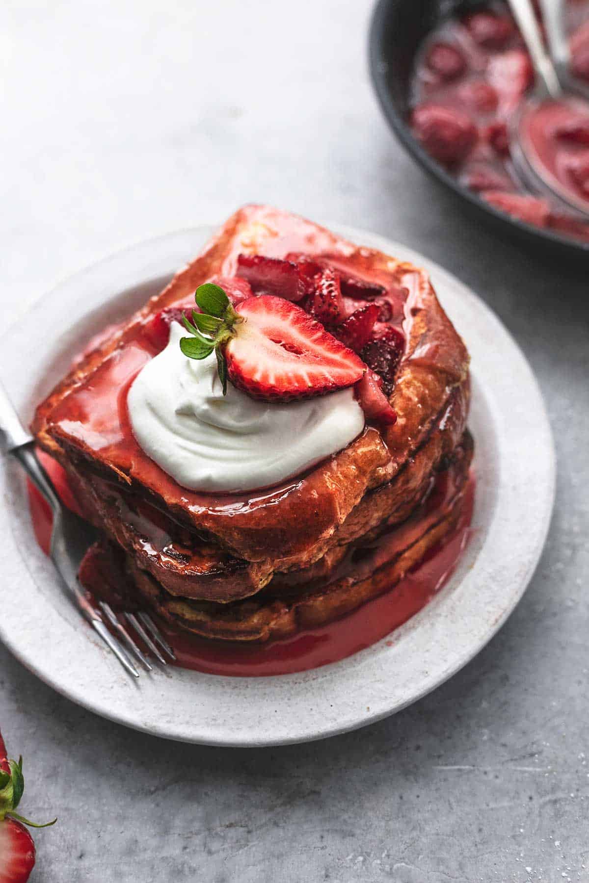 a stack of strawberries and cream French toast on a white plate with a  fork topped with strawberry sauce and whipped cream.