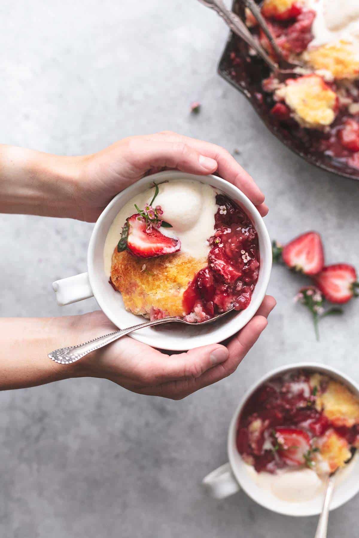 over head view of hands holding bowl of strawberry cobbler with ice cream