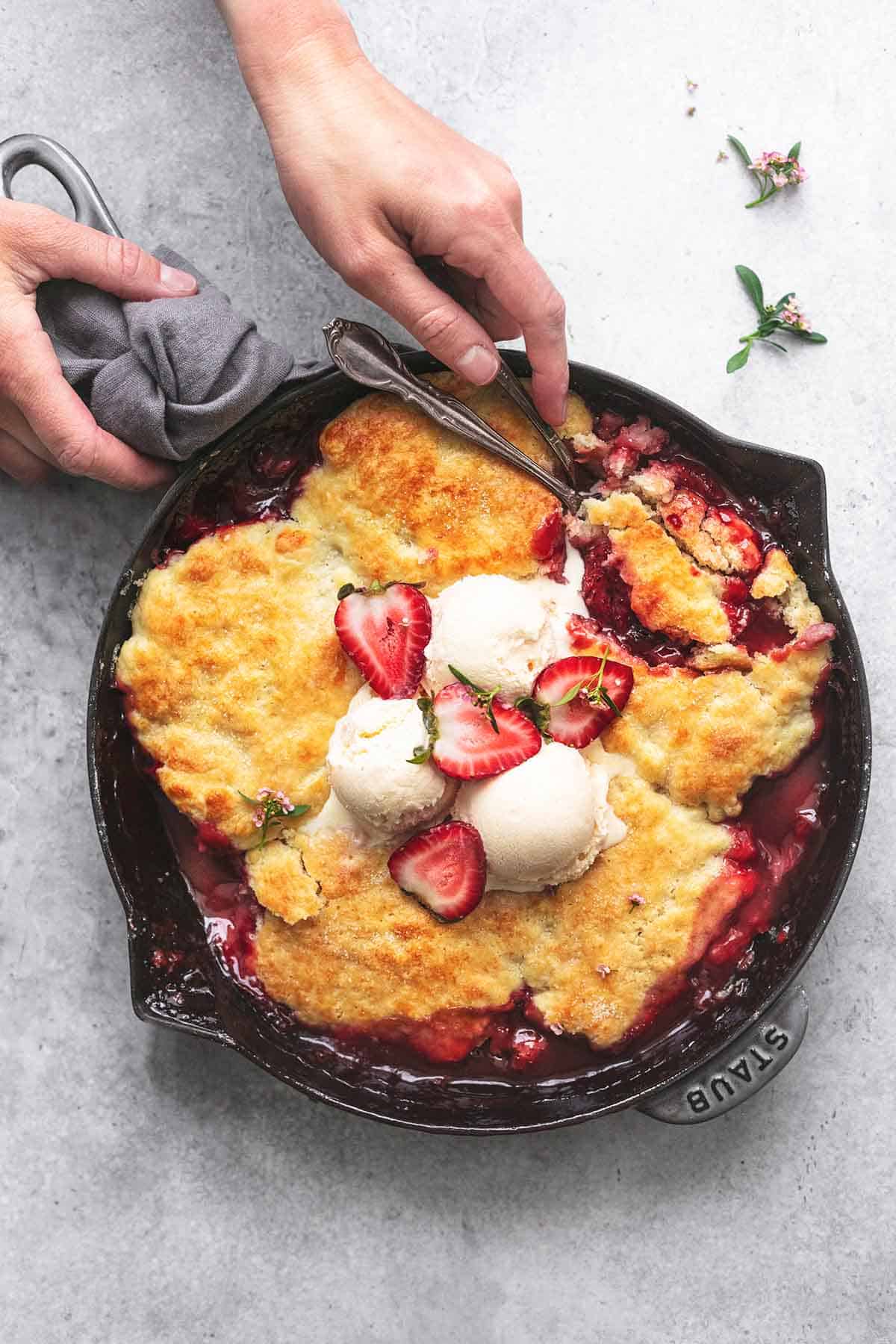 overhead view of hands serving skillet full of baked strawberry cobbler