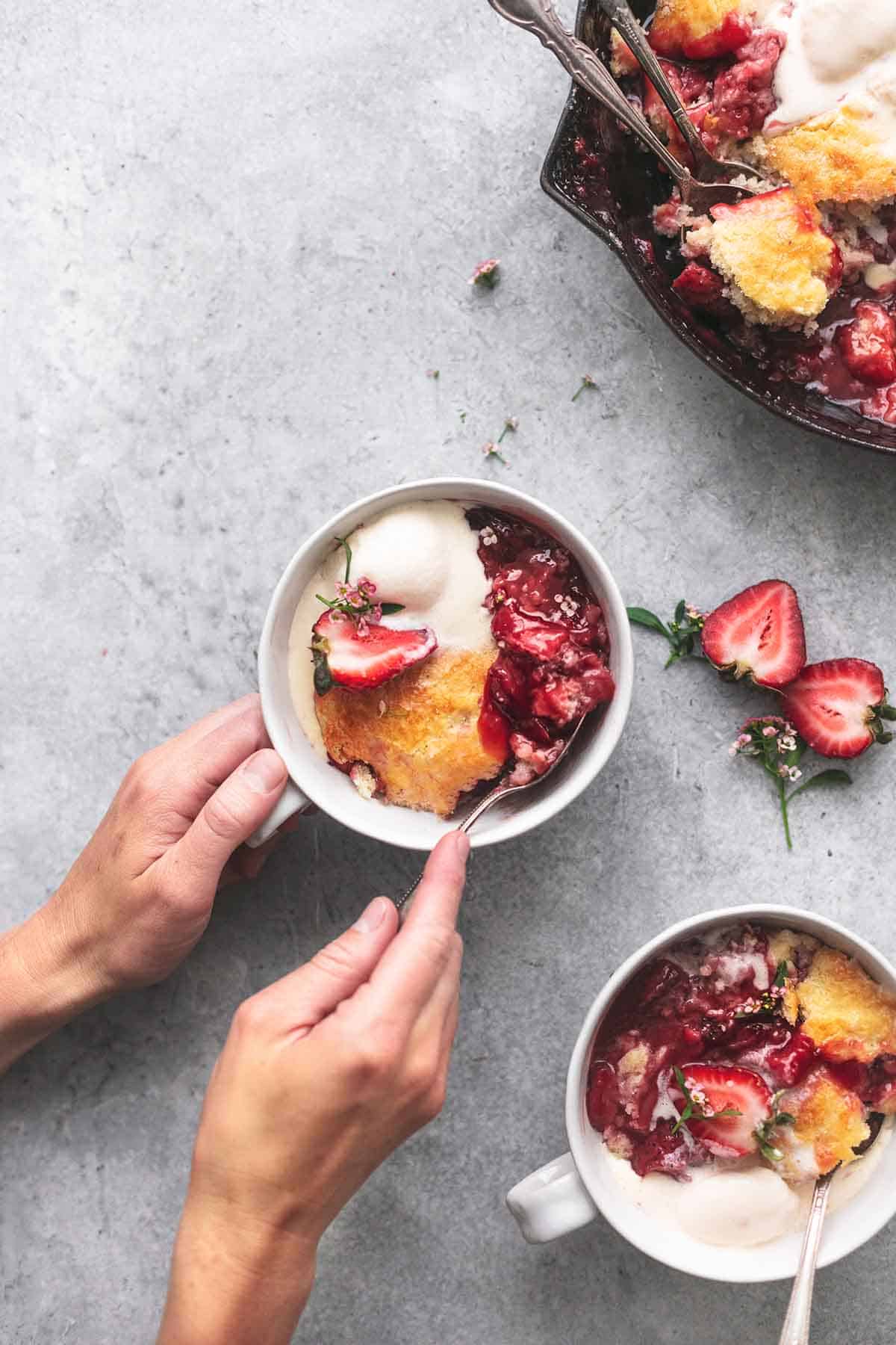 top view of hands dipping a spoon into strawberry cobbler in a bowl with more cobbler in another bowl and cast iron on the side.
