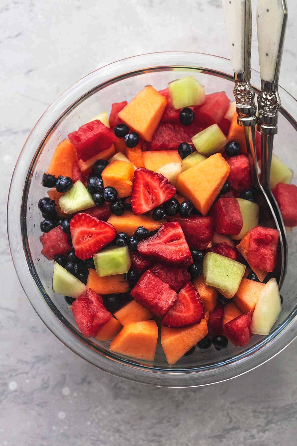 overhead view of fruit salad with berries and melon chunks in a glass bowl with serving spoons