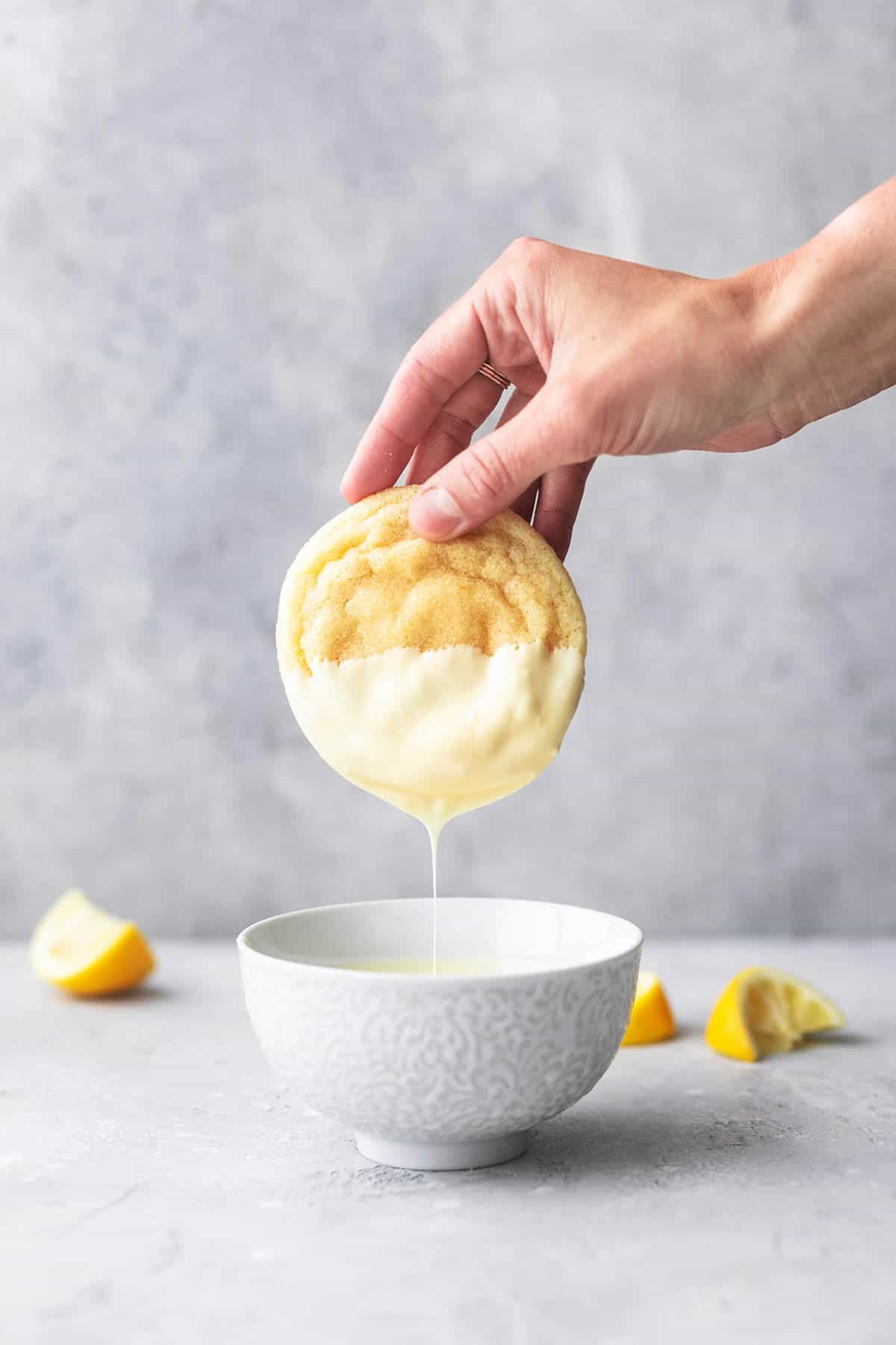 a hand dipping a snow capped lemon cookie into melted white chocolate in a bowl.