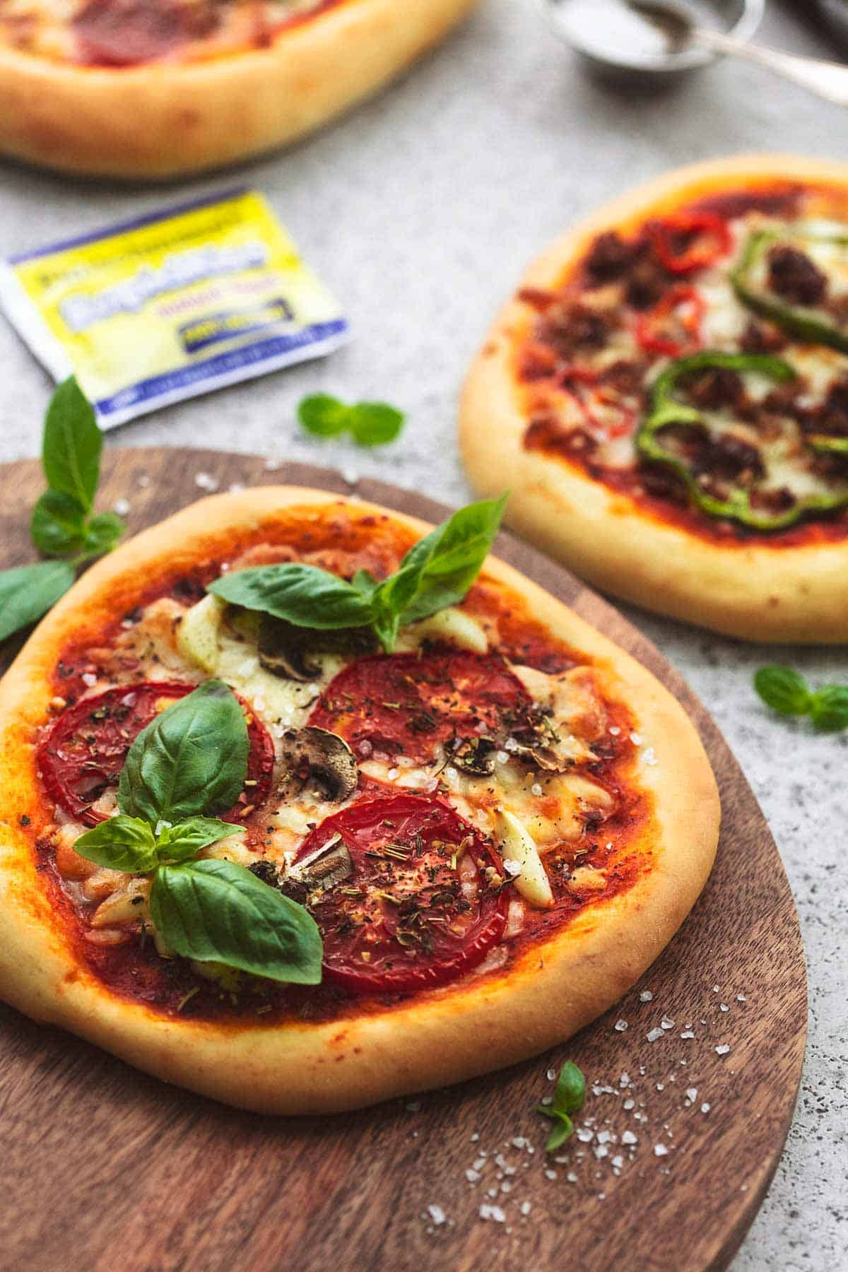 a personal pizza on a wooden cutting board with a yeast packet and more personal pizzas in the background.