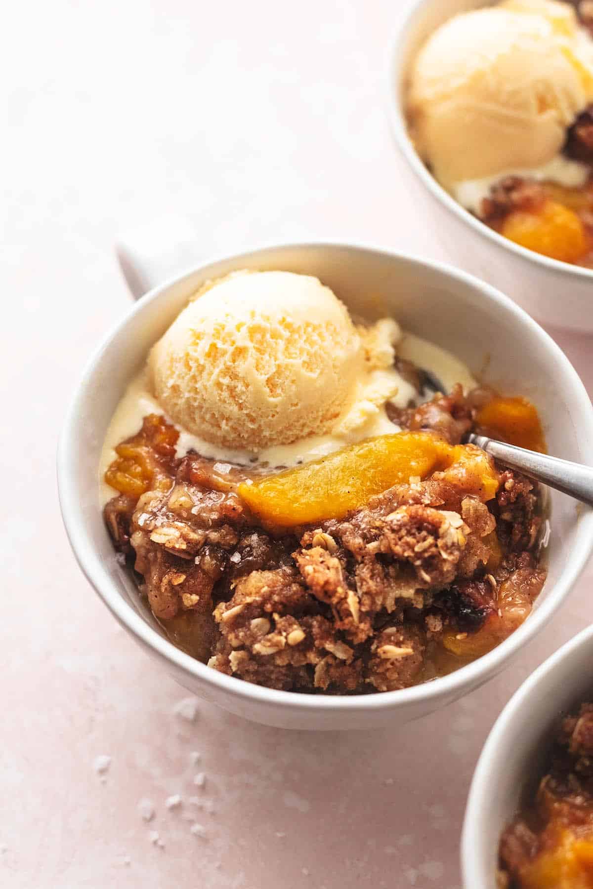 three bowls of peach crisp with spoons and vanilla ice cream