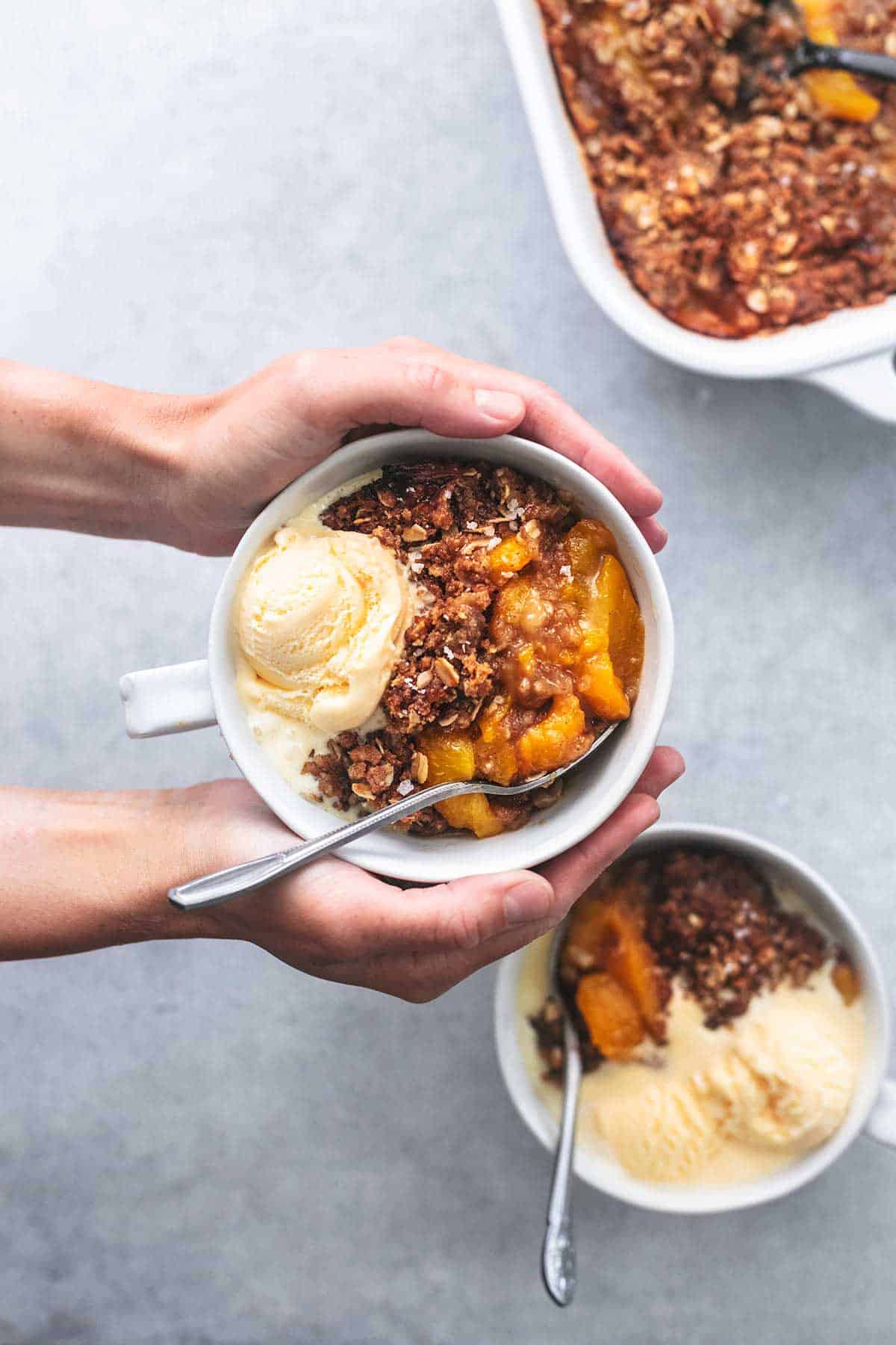 top view of hands holding a bowl of peach crisp with another bowl and a baking pan of peach crisp on the side.