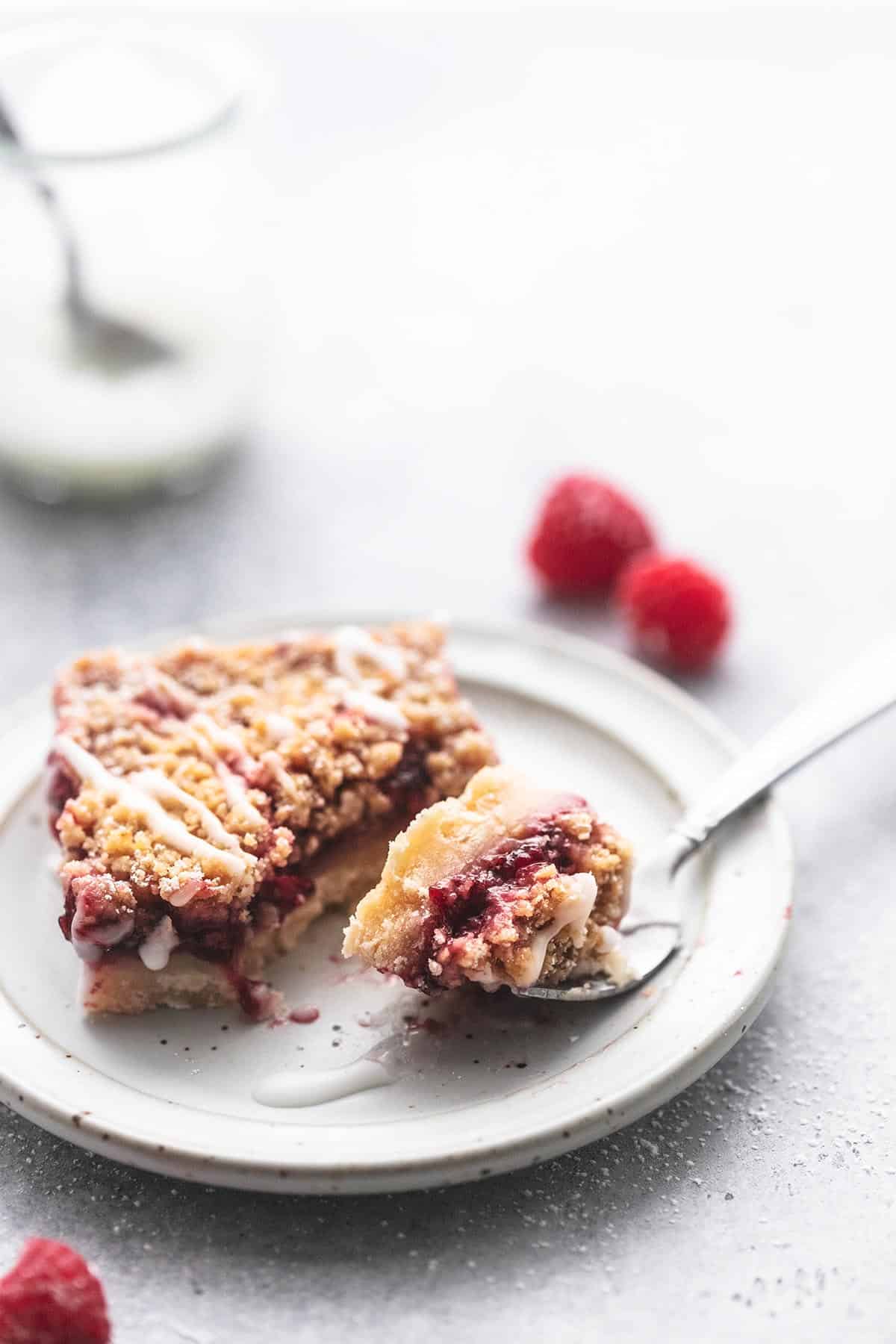 a raspberry crumb bar on a plate with a bite on a fork with a jar of glaze and raspberries on a table.