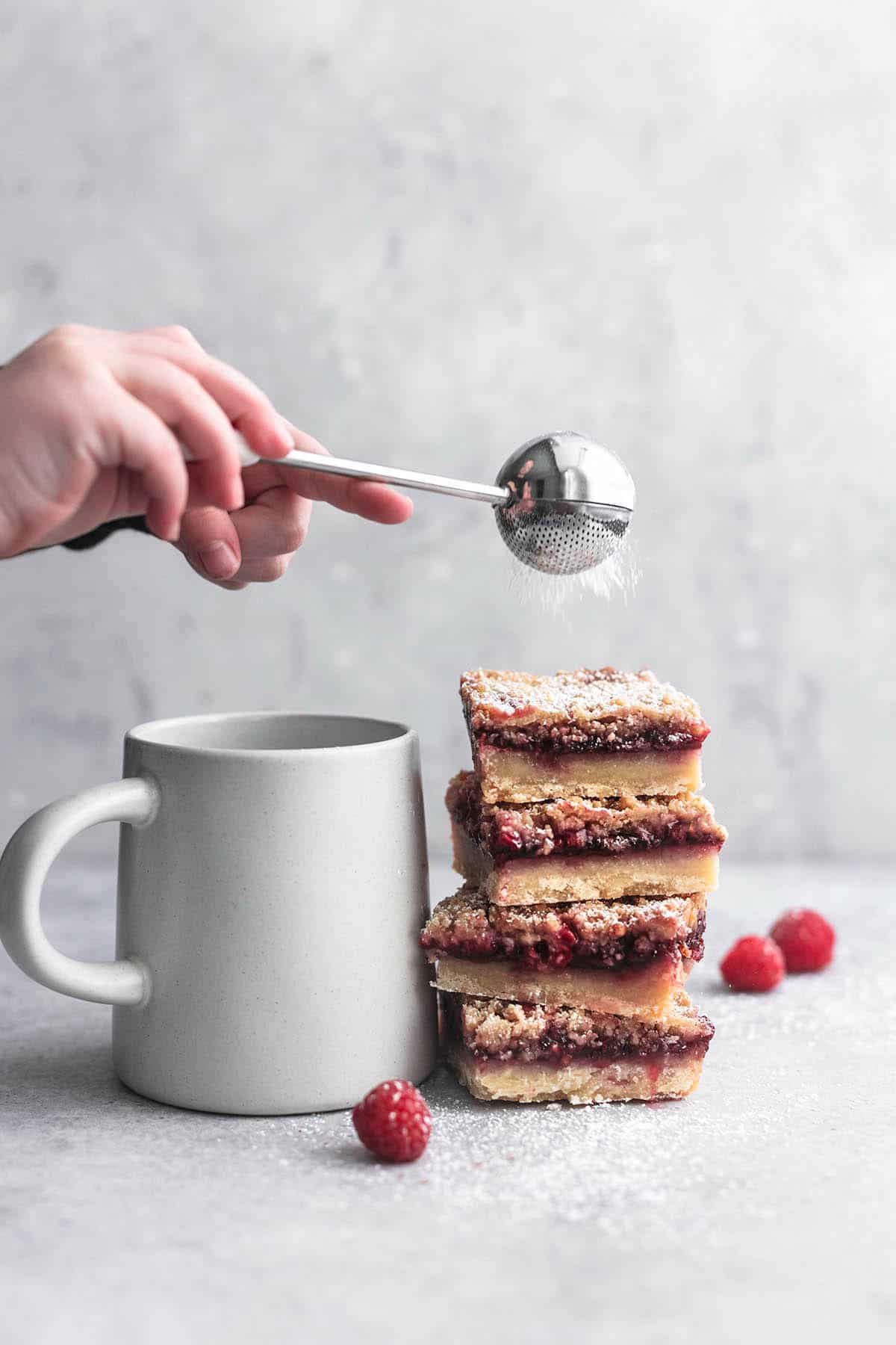 a hand holding a powdered sugar shaker over a stack of raspberry crumb bars.