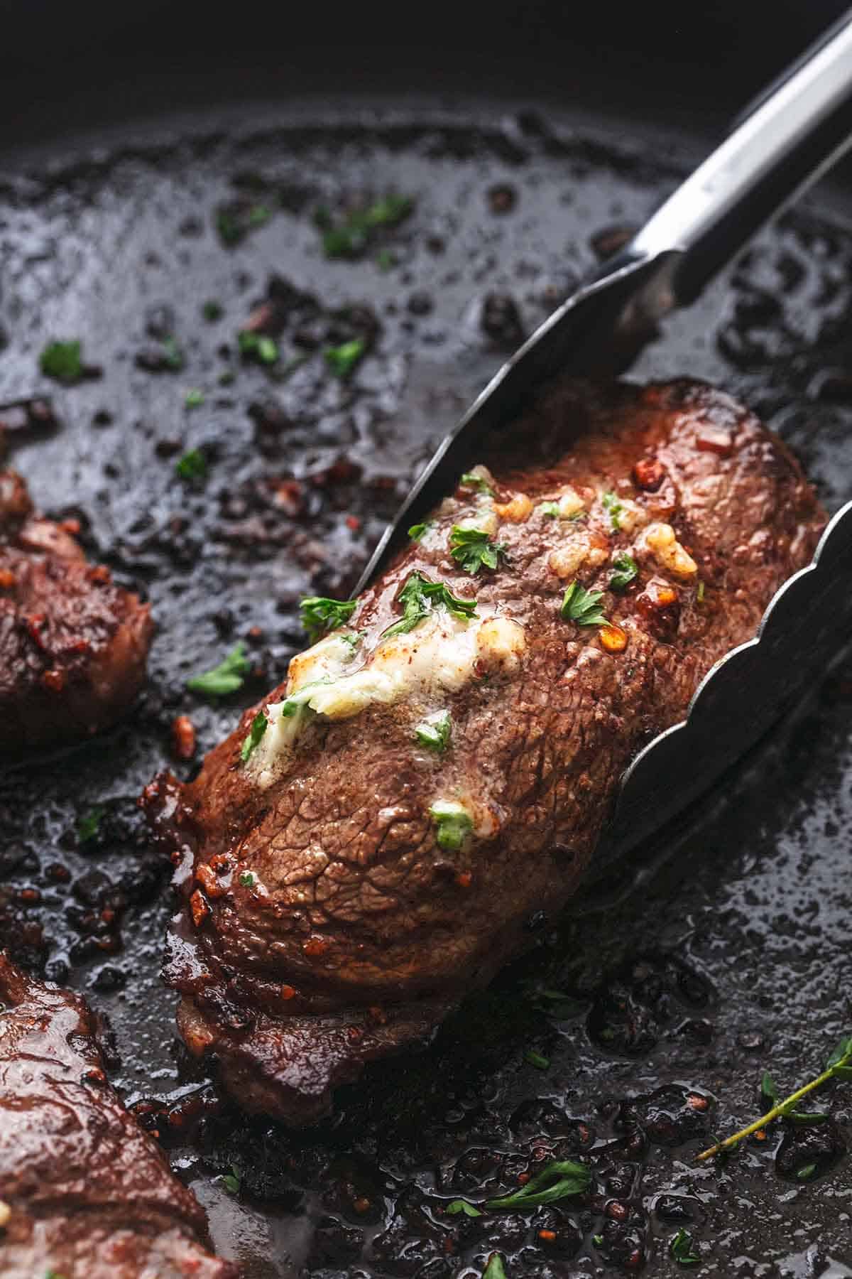 close up of tongs holding a sirloin steak with butter on top.