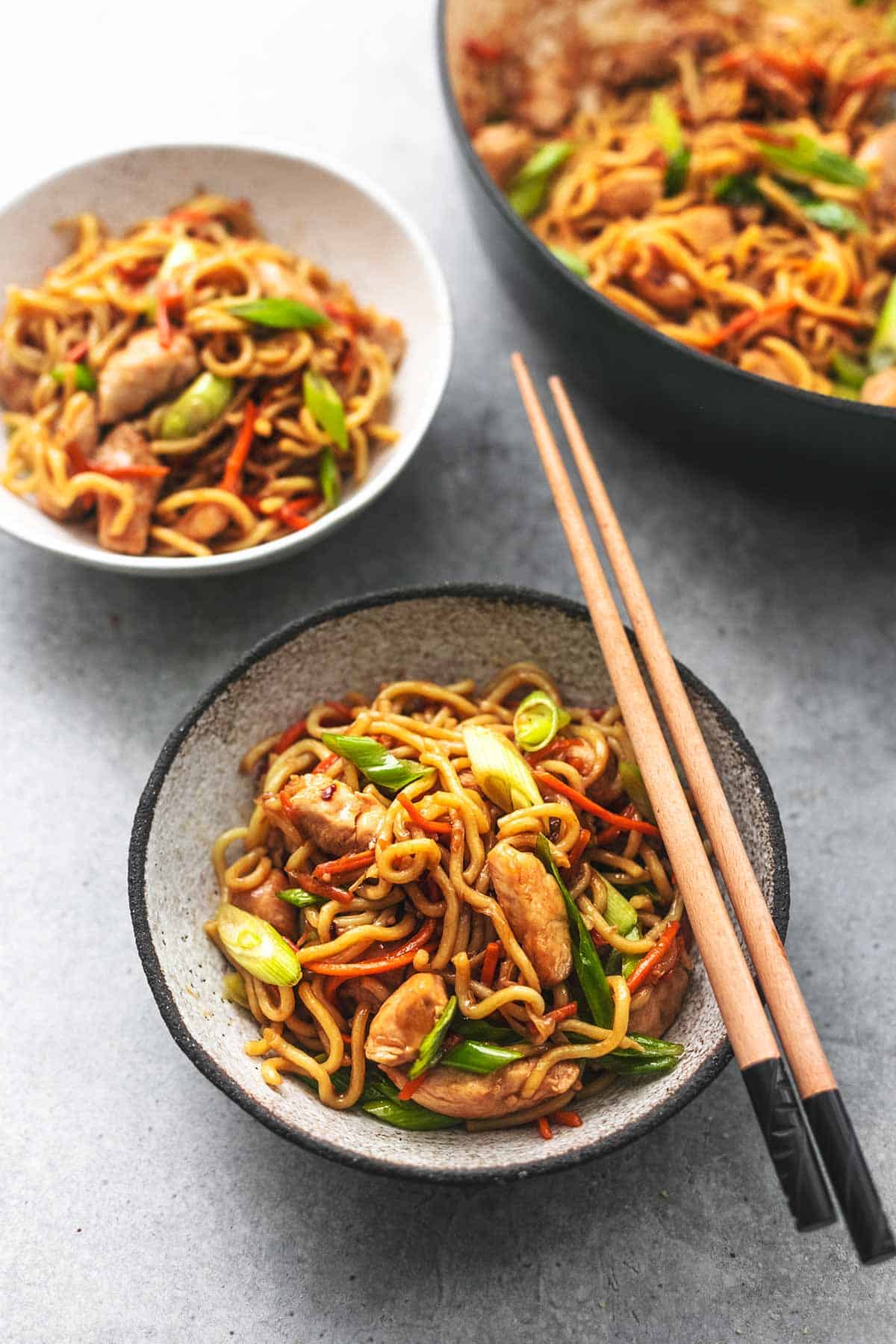 a bowl of chicken chow mein with chopsticks on top with a mother bowl and skillet on the side.