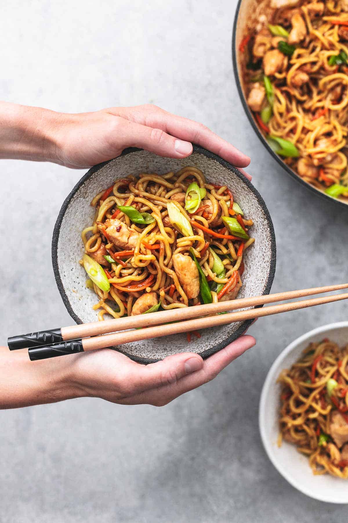 top view of hands holding a bowl of chicken chow mein above another bowl and skillet.