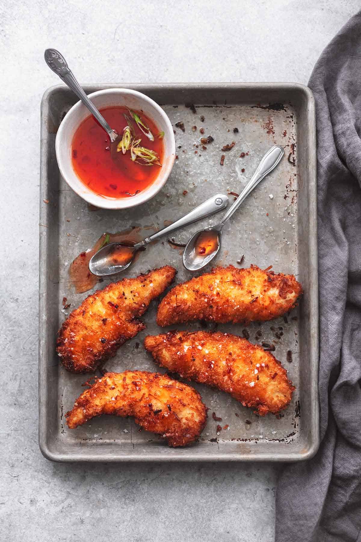 top view of coconut chicken tenders on a baking sheet with two spoons and a bowl of red sauce on the side.