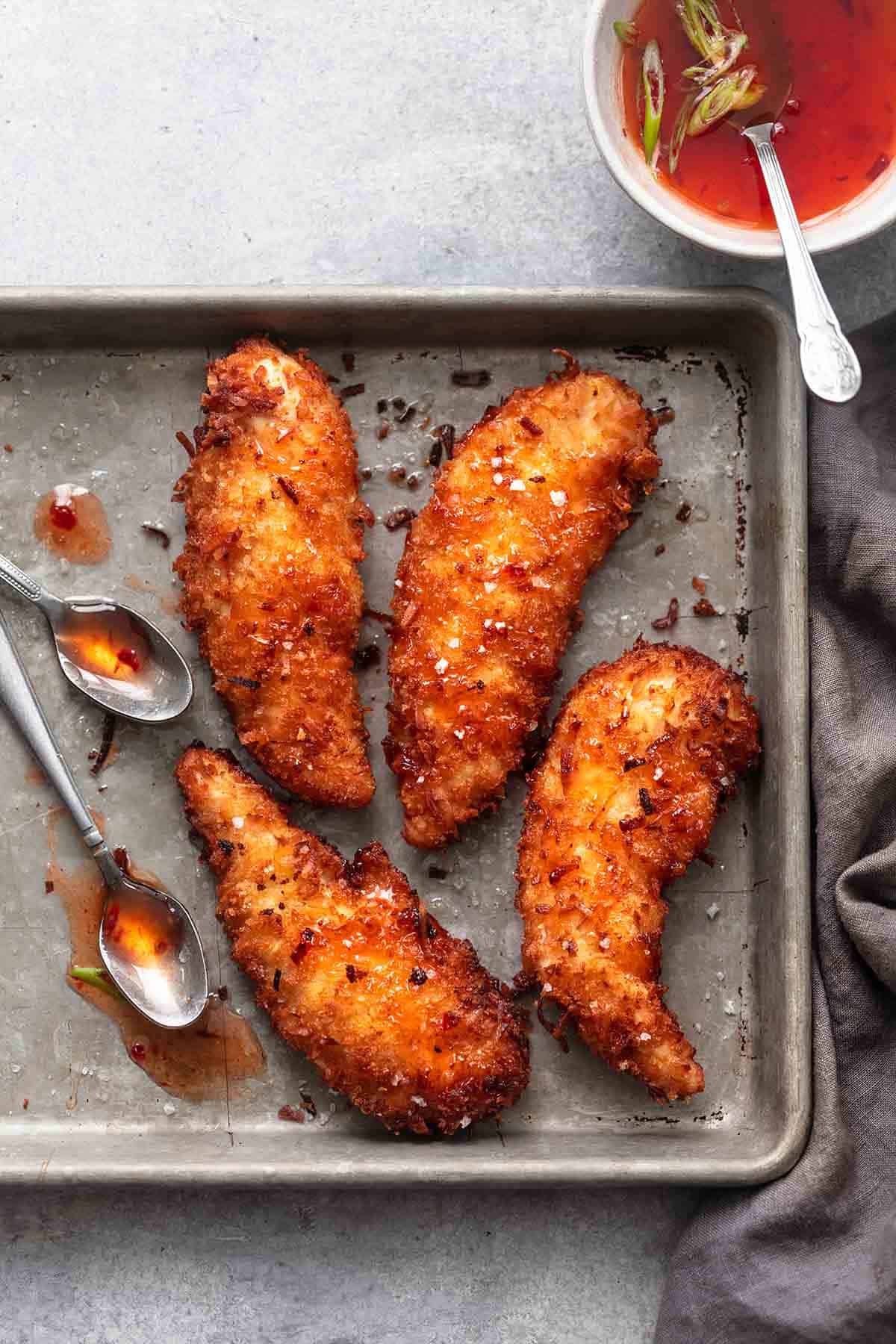 close up top view of coconut chicken tenders with two spoons on a baking sheet with a bowl of sauce on the side.