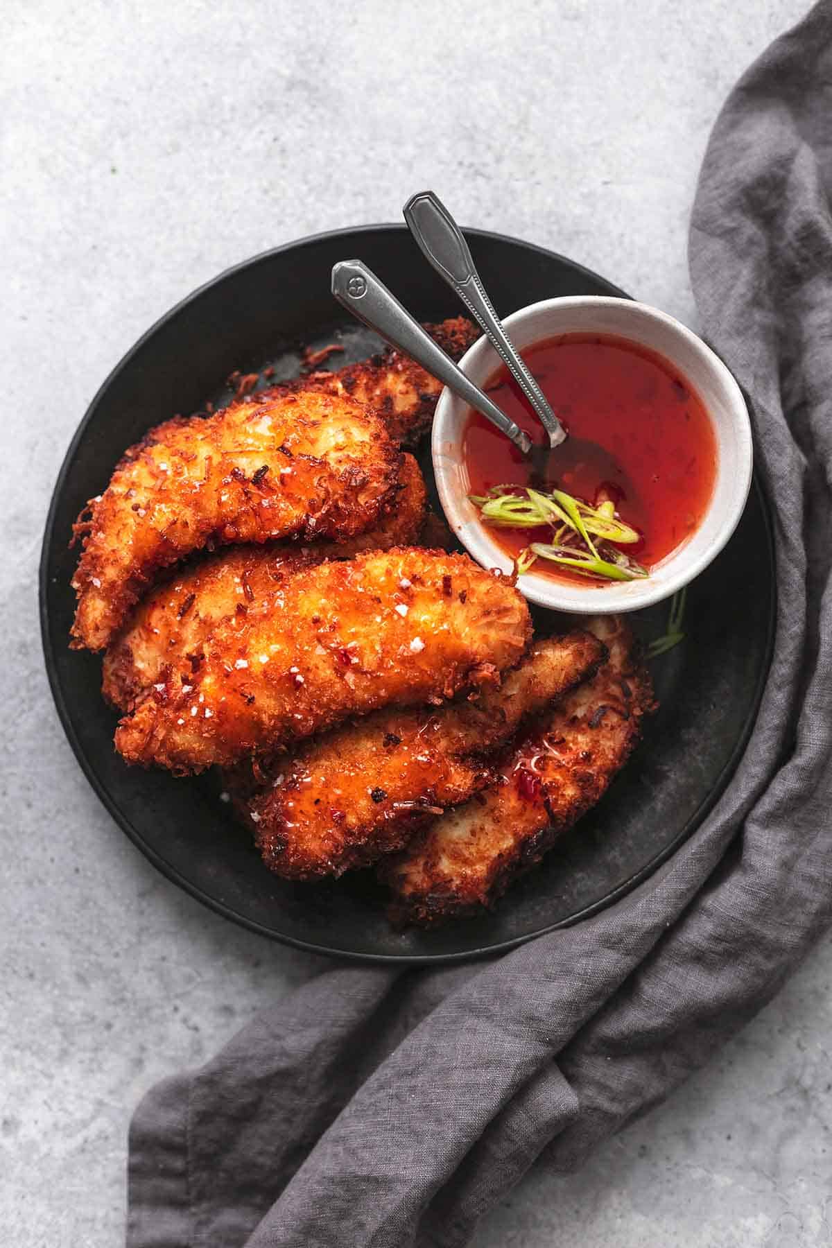 top view of coconut chicken tenders on a black plate with a bowl of sauce on the side.