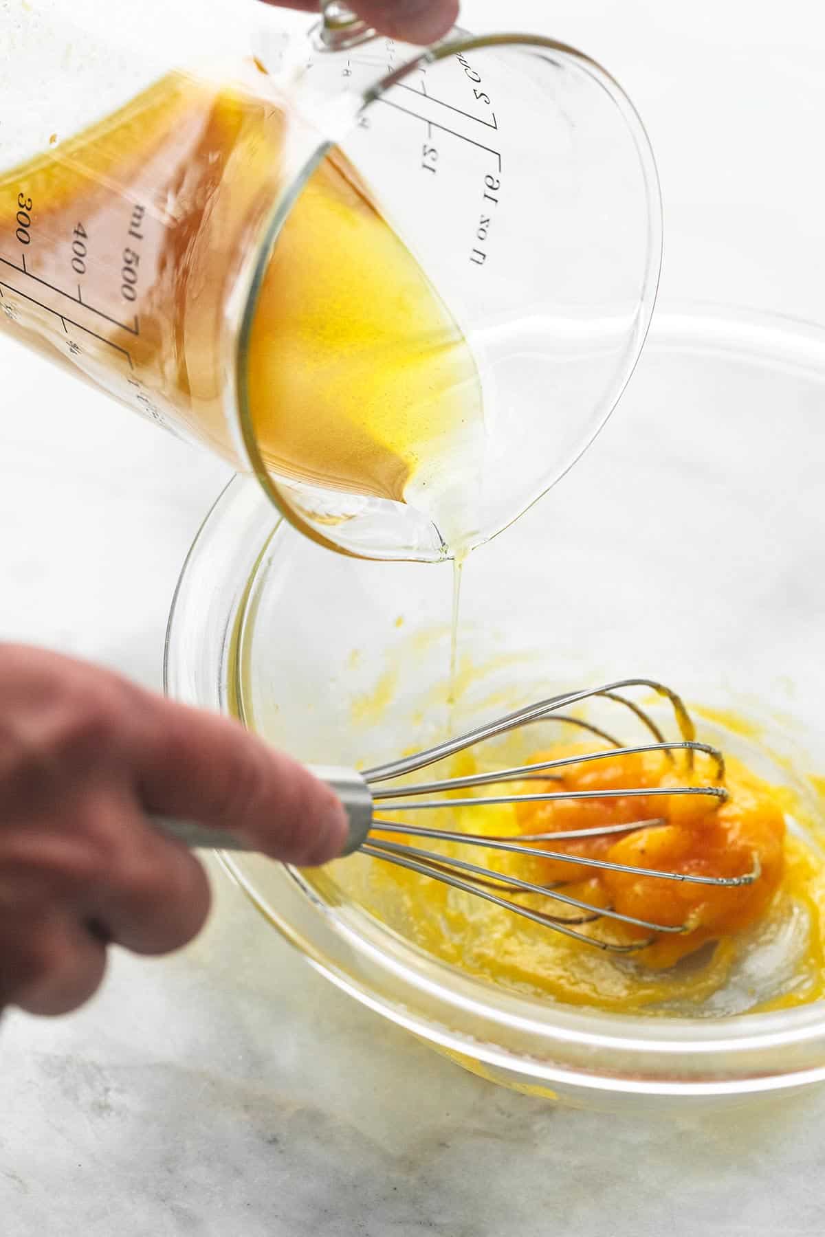hands whisking egg yolks in a glass bowl and pouring olive oil into the bowl.