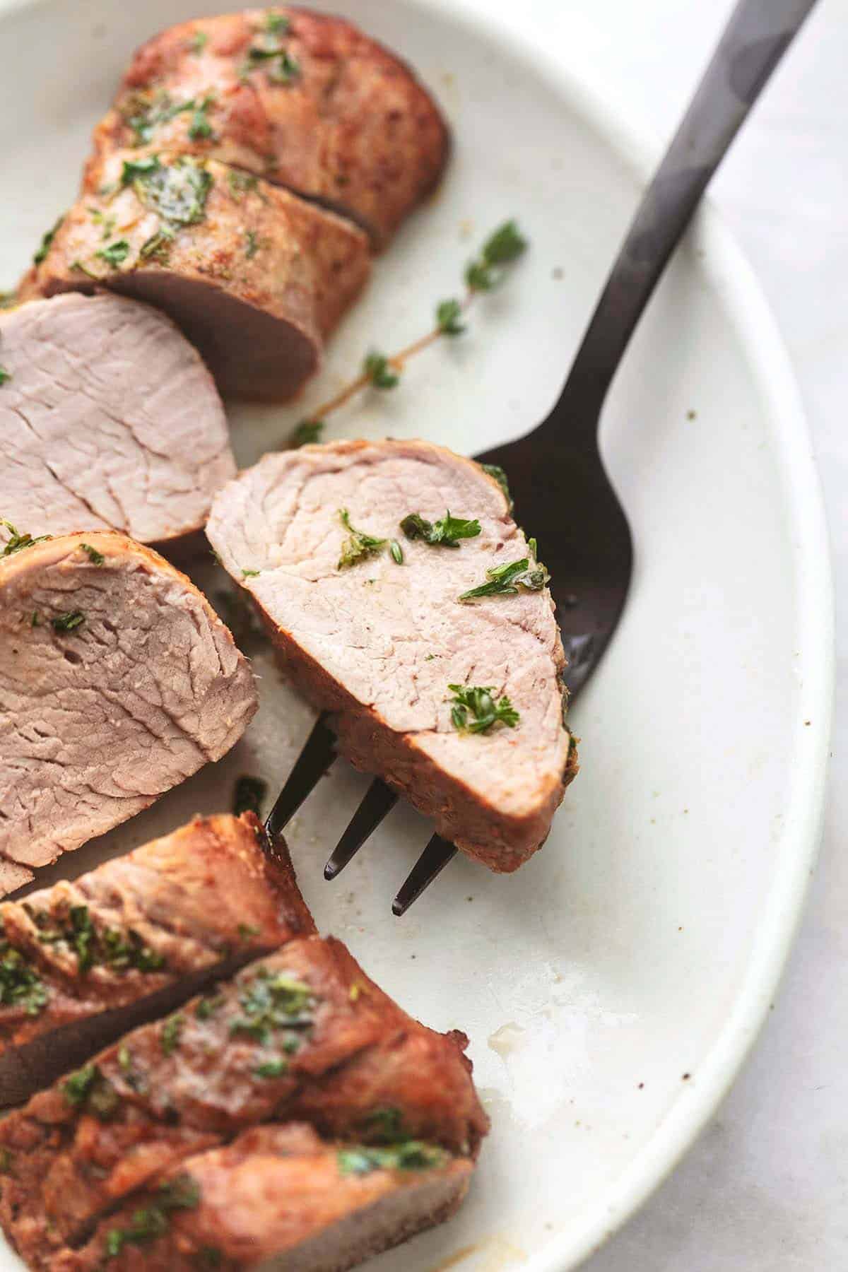 close up of a slice of garlic butter roasted pork tenderloin with herbs on a serving fork with the rest of the tenderloin on the side both on a platter.