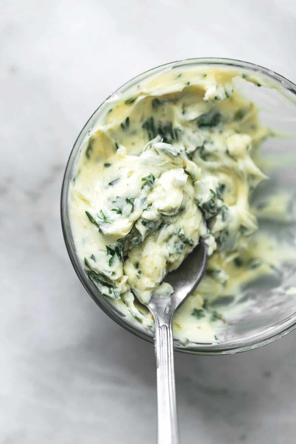 close up top view of a small glass bowl of garlic herb butter with a spoon.