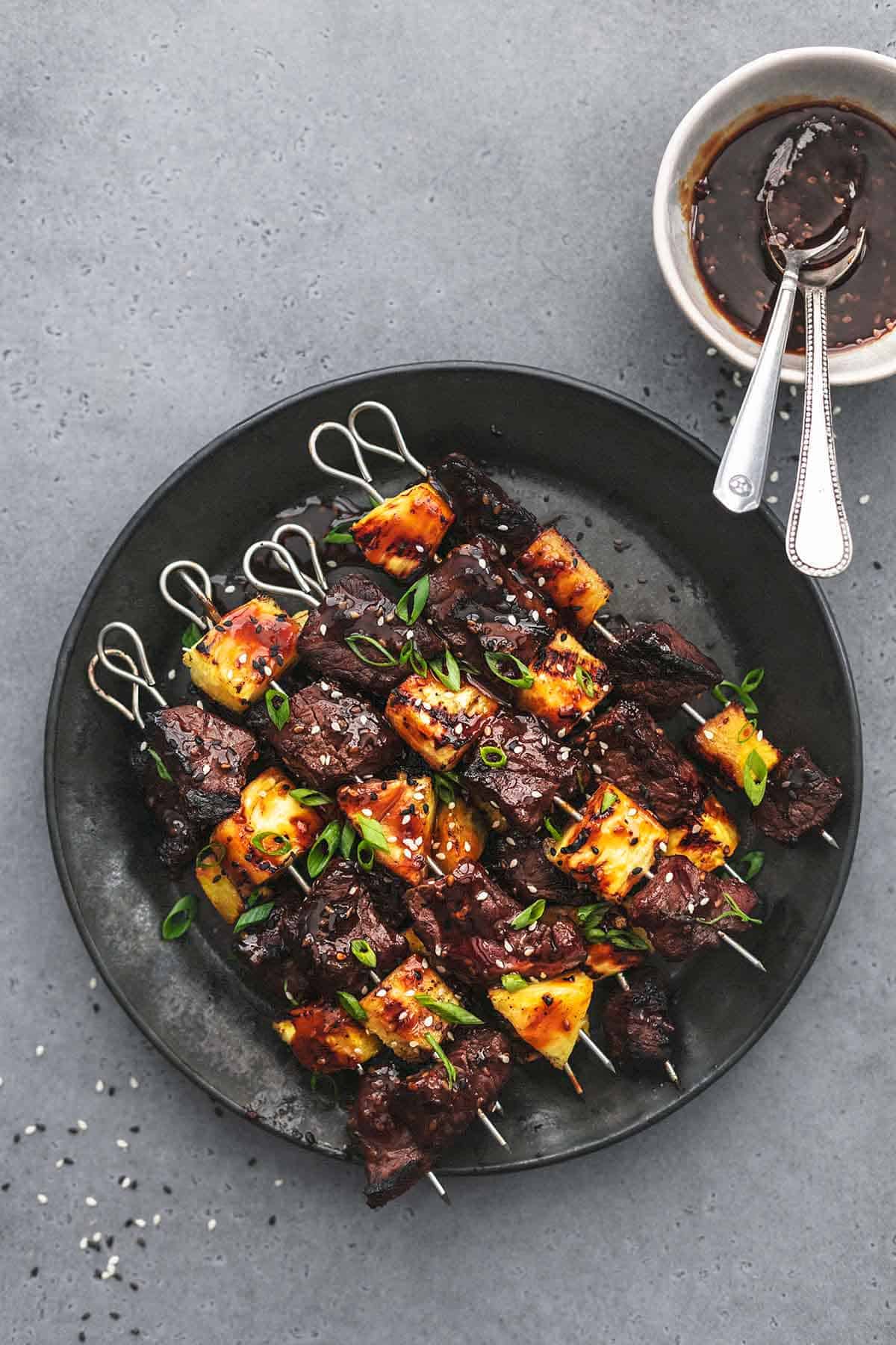 top view of teriyaki steak and pineapple kabobs on a plate with a small bowl with sauce and spoons on the side.