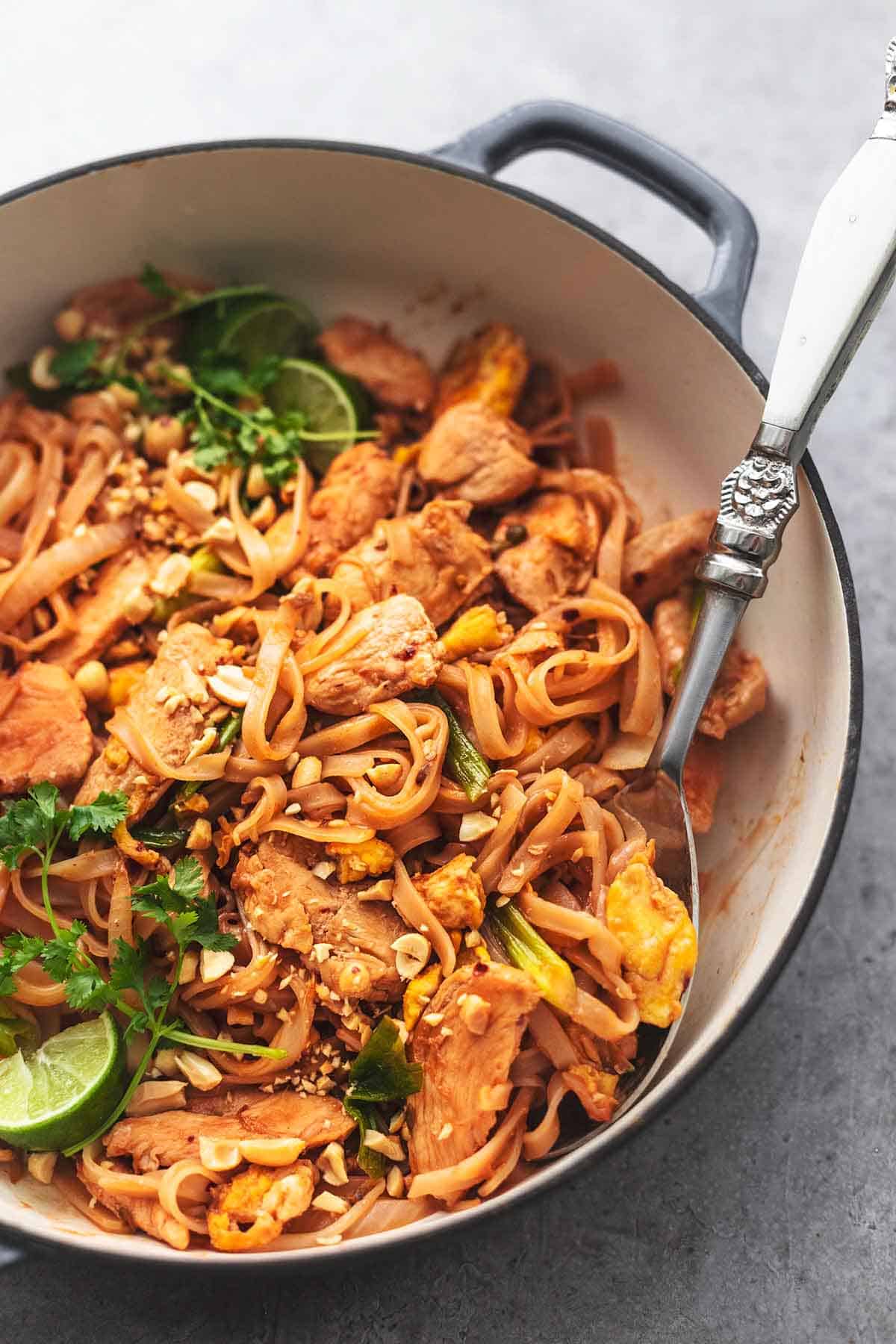 a serving spoon in a skillet filled with chicken and flat noodles with eggs and limes