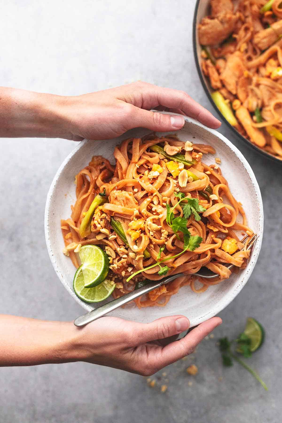 hands holding a plate of chinese noodles above a skillet filled with more noodles