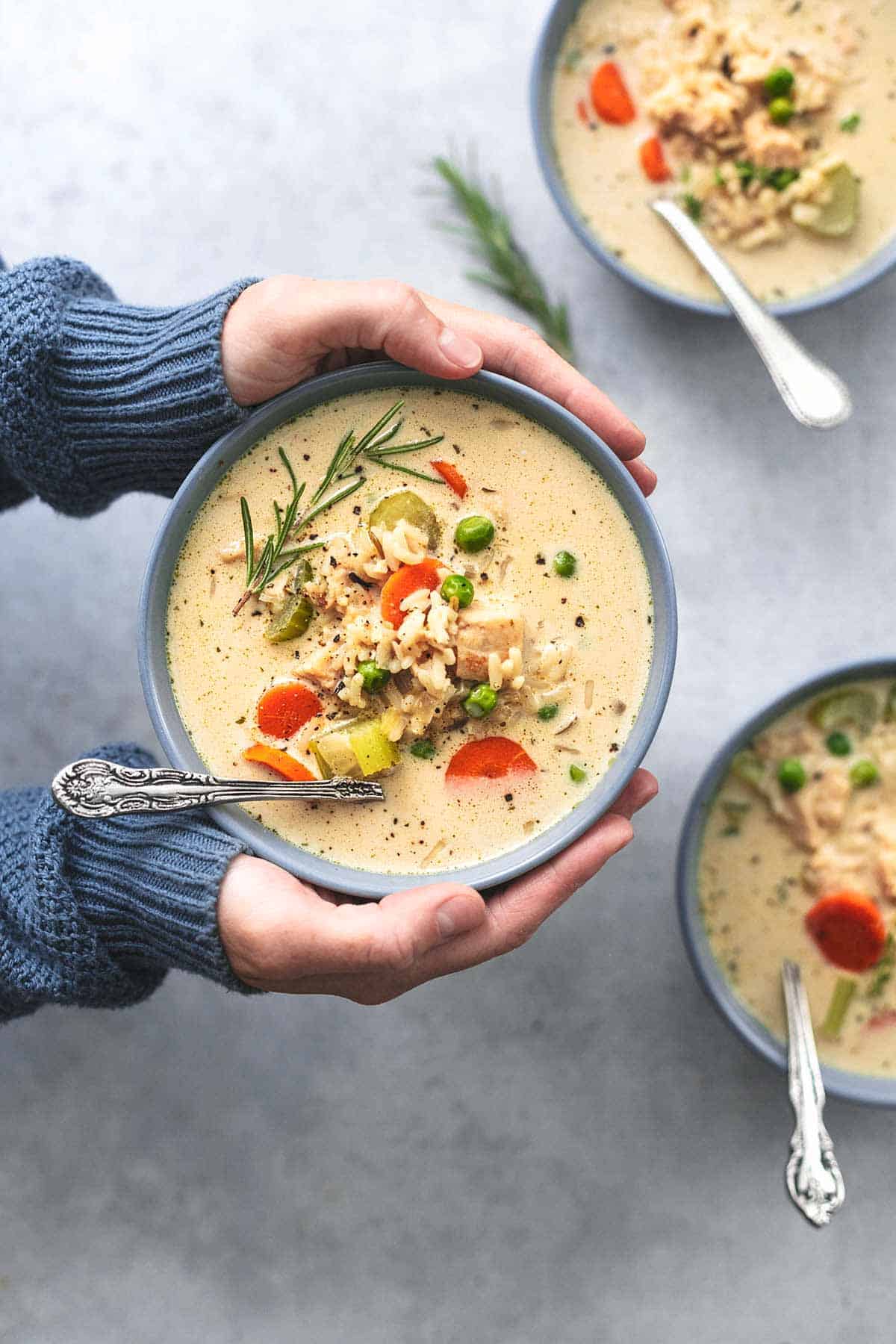 top view of hands holding a bowl of chicken and rice soup over two more bowls of soup.