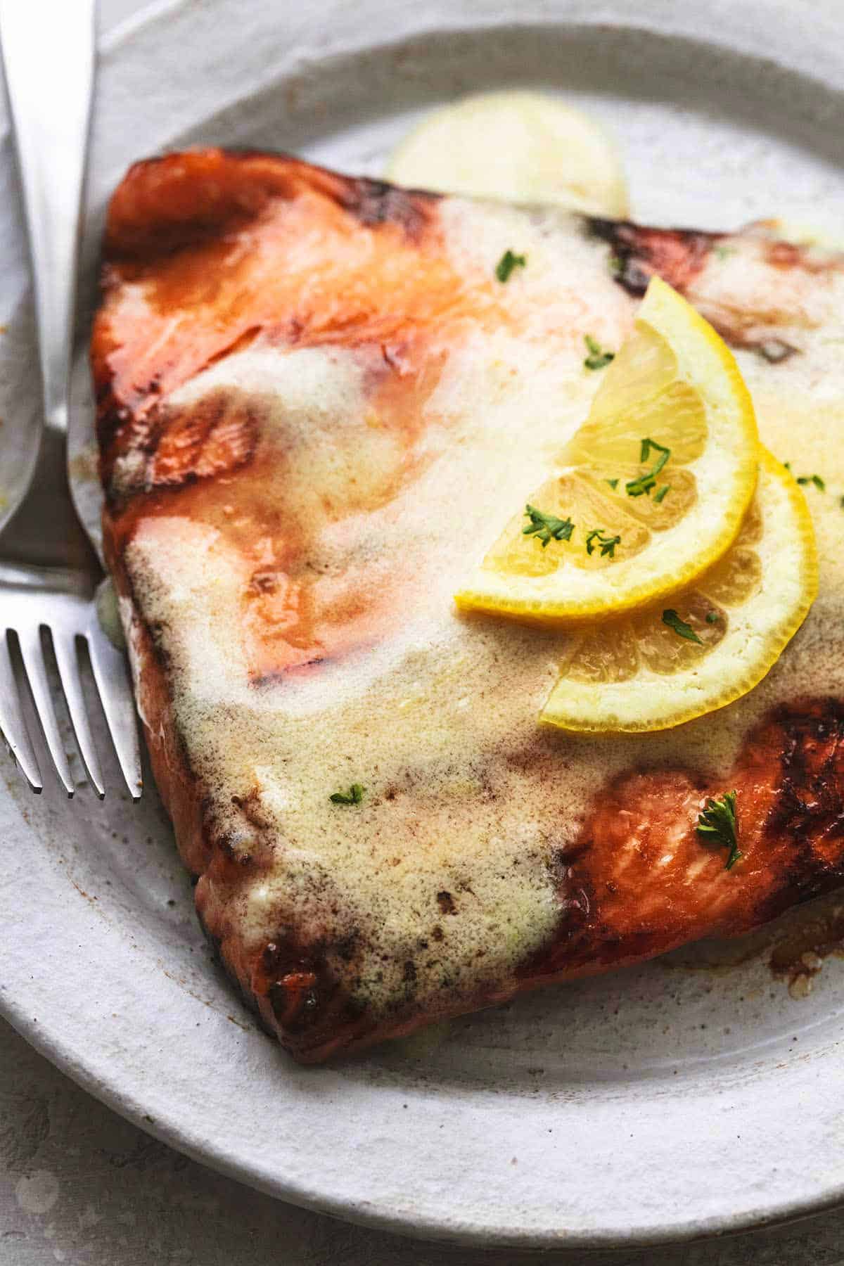 close up of cooked salmon with lemon butter sauce and lemon slices on top with a fork on the side on a plate.