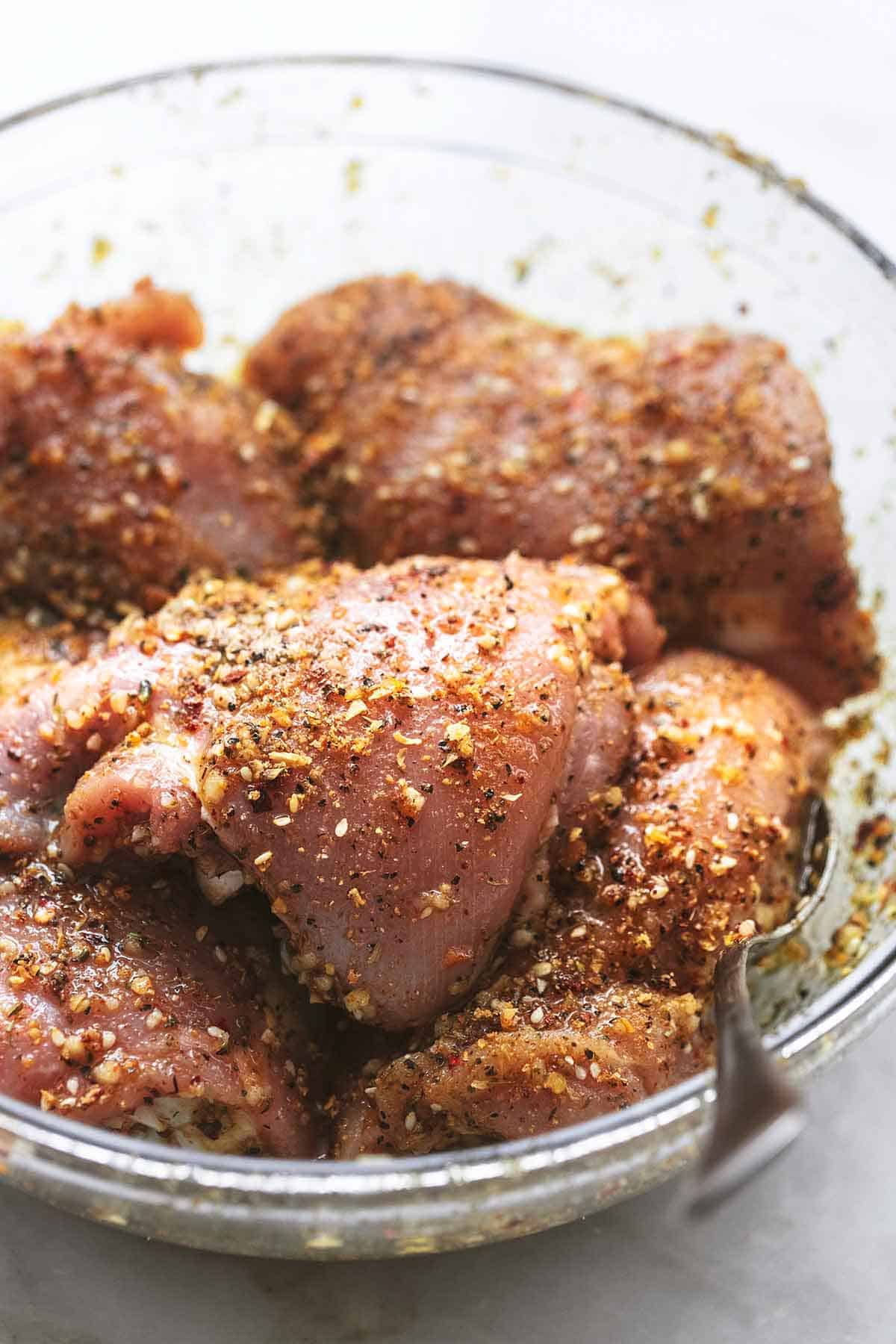 close up of uncooked chicken thighs with spices in a clear glass bowl.
