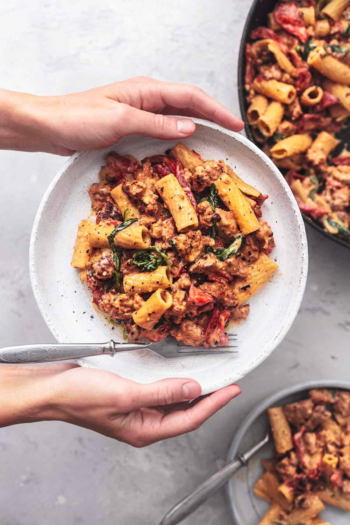 hands holding plate of pasta above a skillet and second plate of pasta