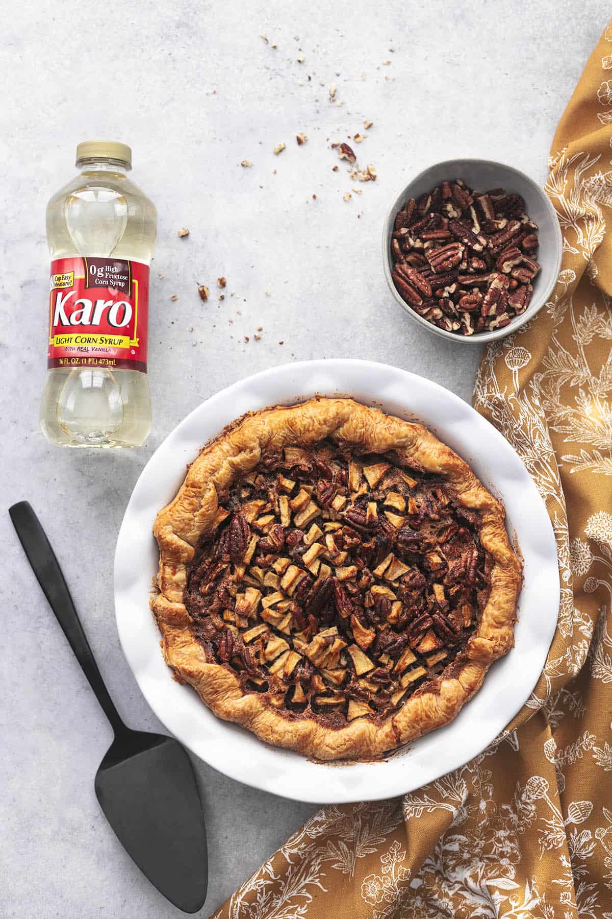 top view of apple pecan pie in a dish with a cup of chopped pecans, a yellow floral linen and a Karo corn syrup bottle.