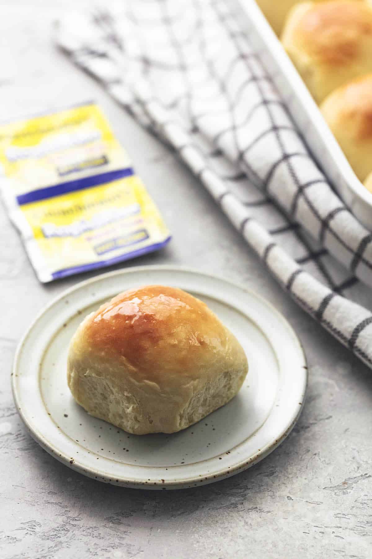 a buttermilk dinner roll on a white plate with yeast packets and baking pan of rolls in background.