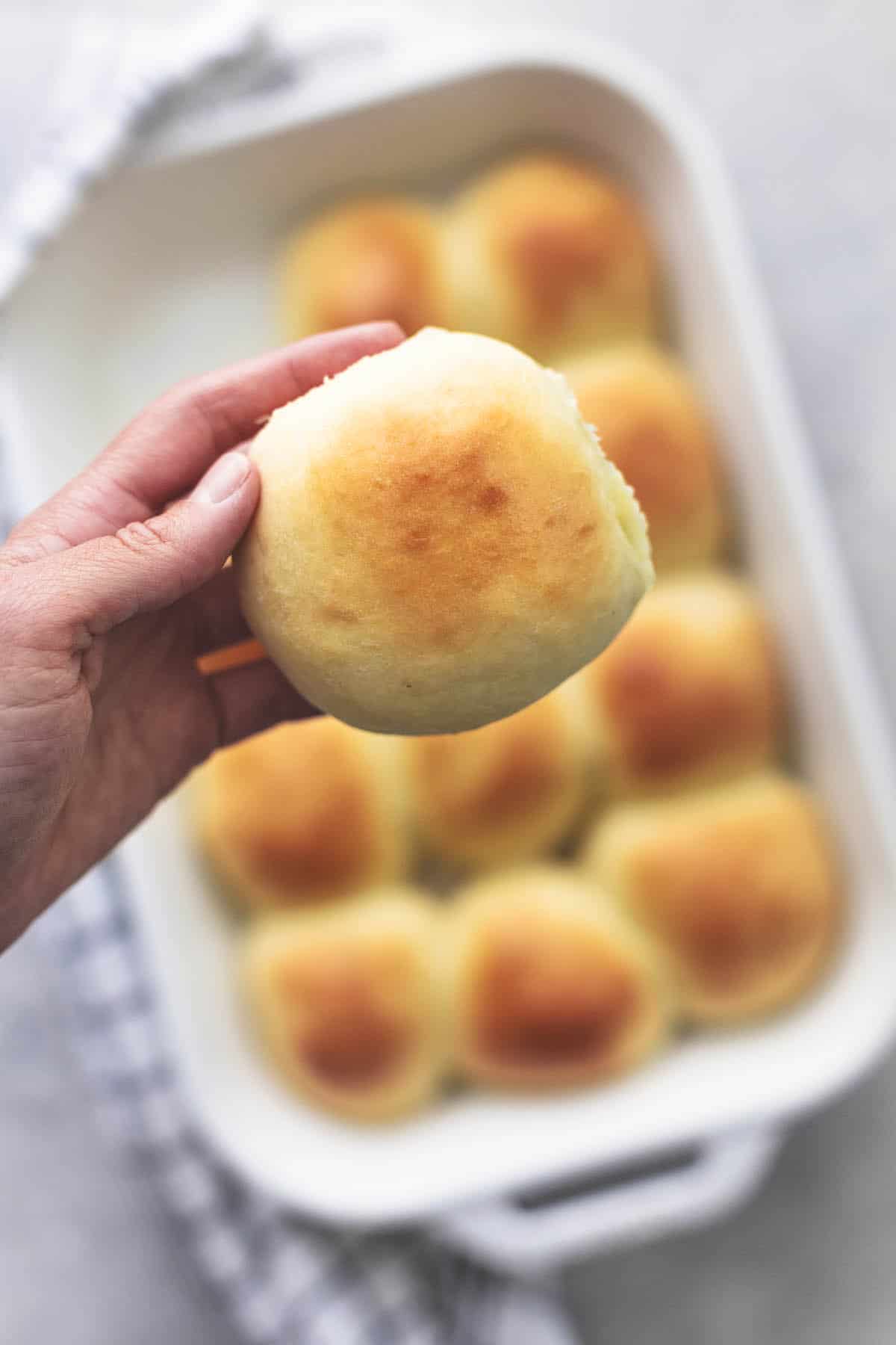top view of a hand holding up a buttermilk dinner roll above more rolls in a baking pan.