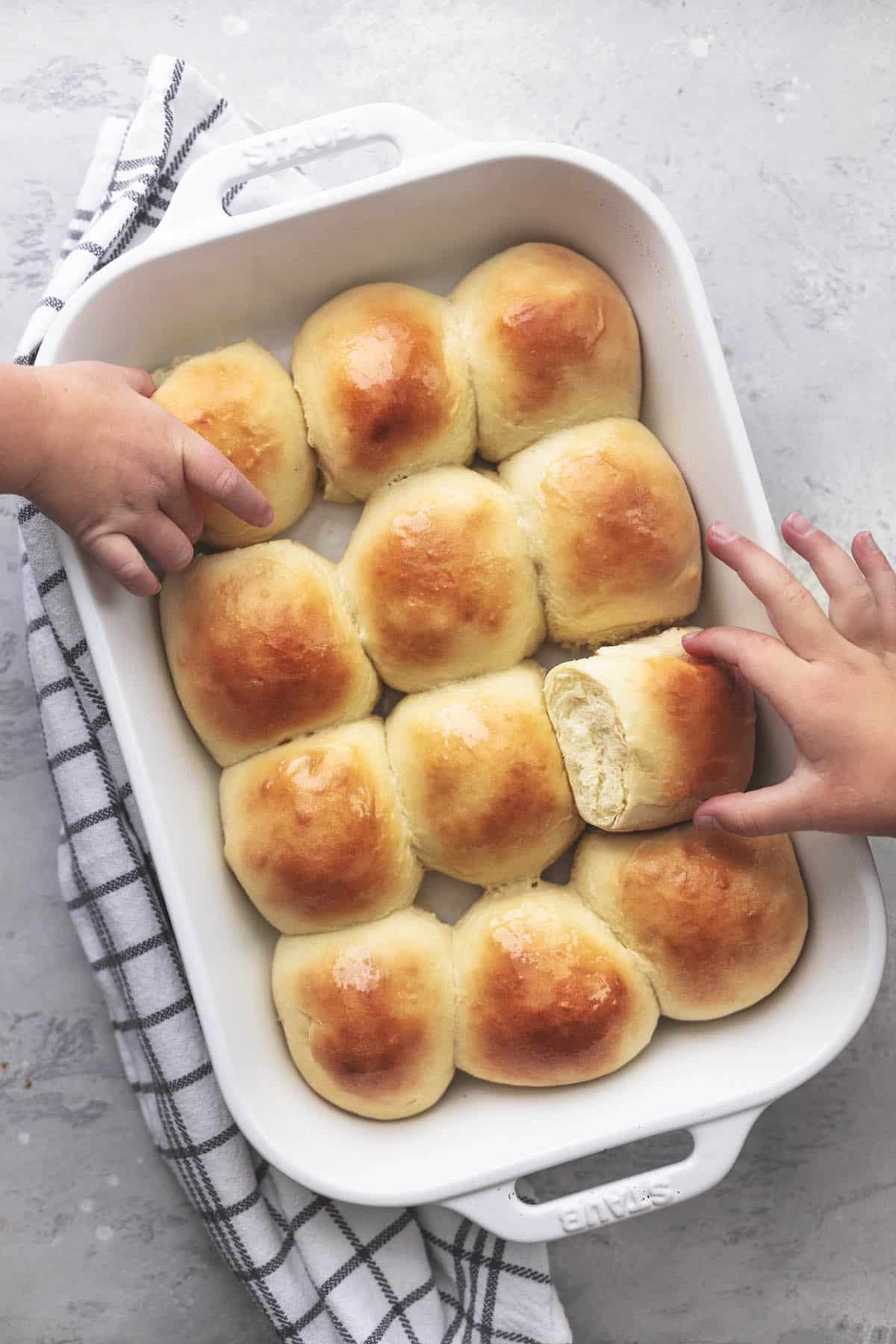 top view of two kid's hands reaching to take buttermilk dinner rolls out of a white baking pan.