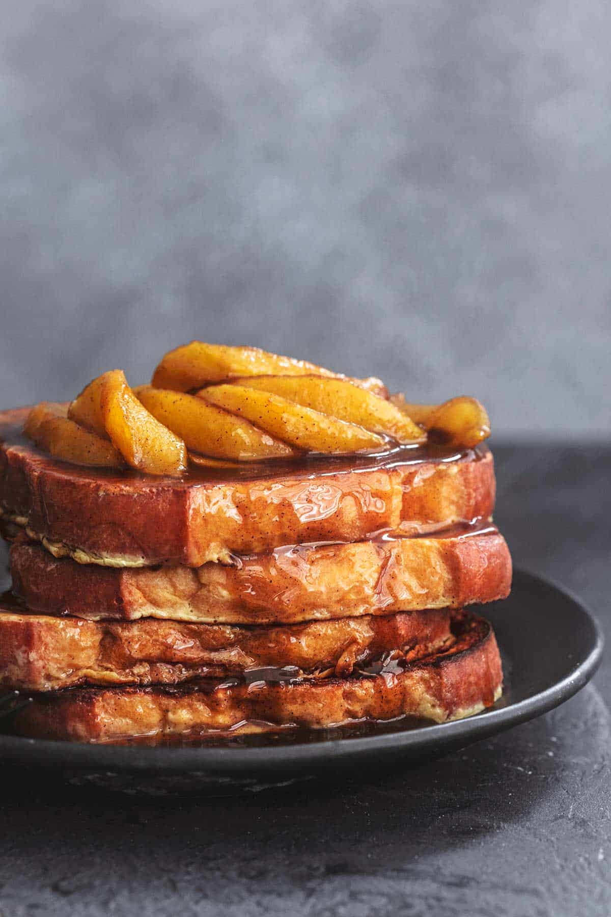 a stack of apple cinnamon French toast topped with sauce and apple slices on a black plate.