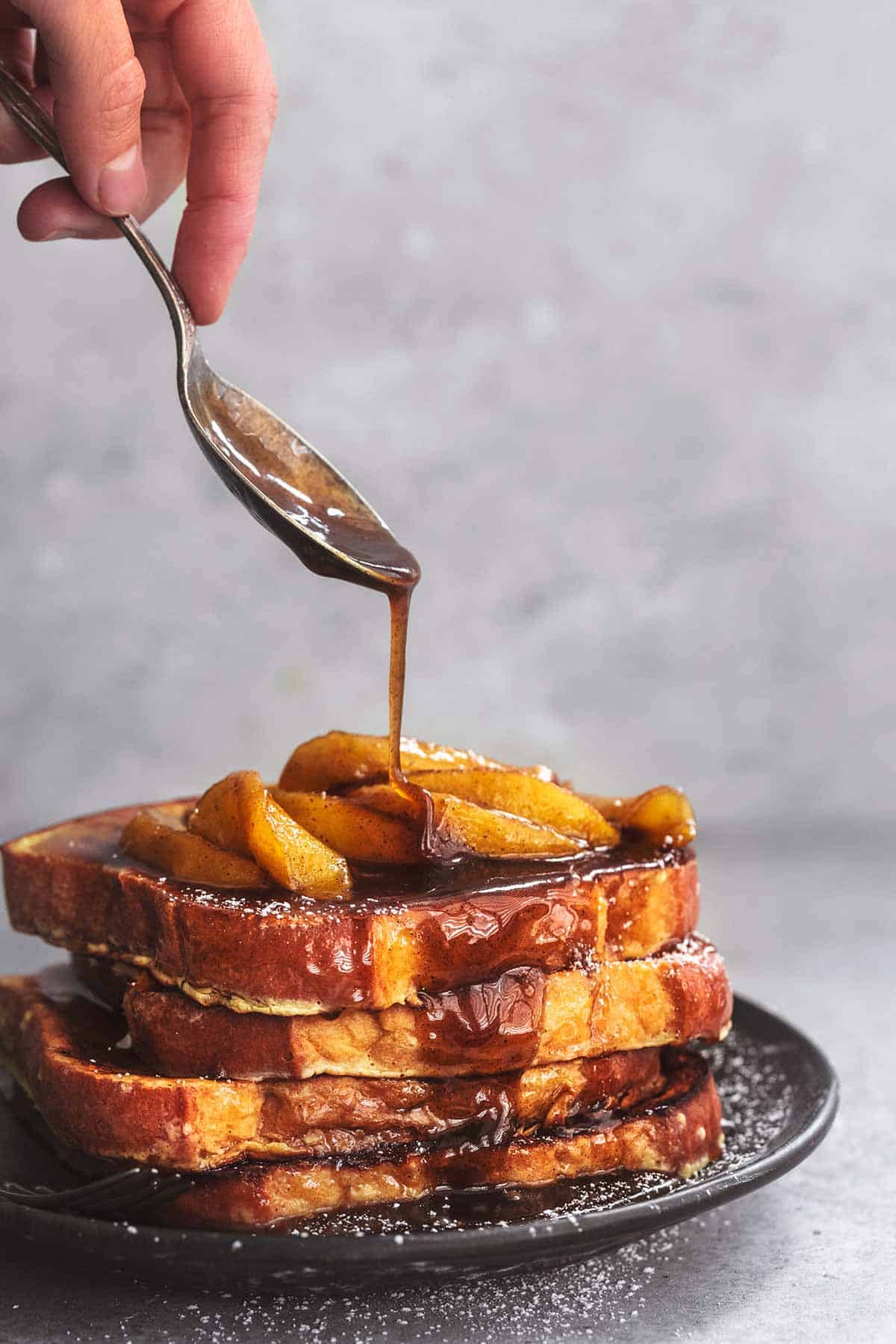 a hand drizzling syrup onto a stack of apple cinnamon French toast on a plate with a spoon.