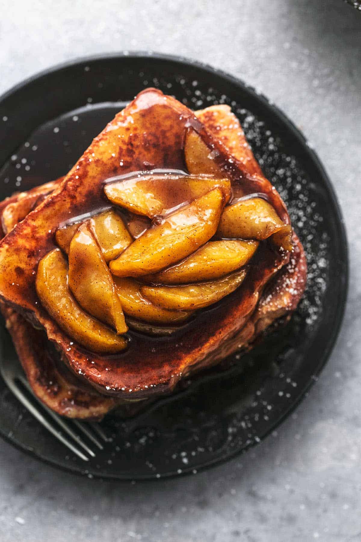 close up top view of a stack of apple cinnamon french toast topped with apple slices and sauce on a black plate with a fork.