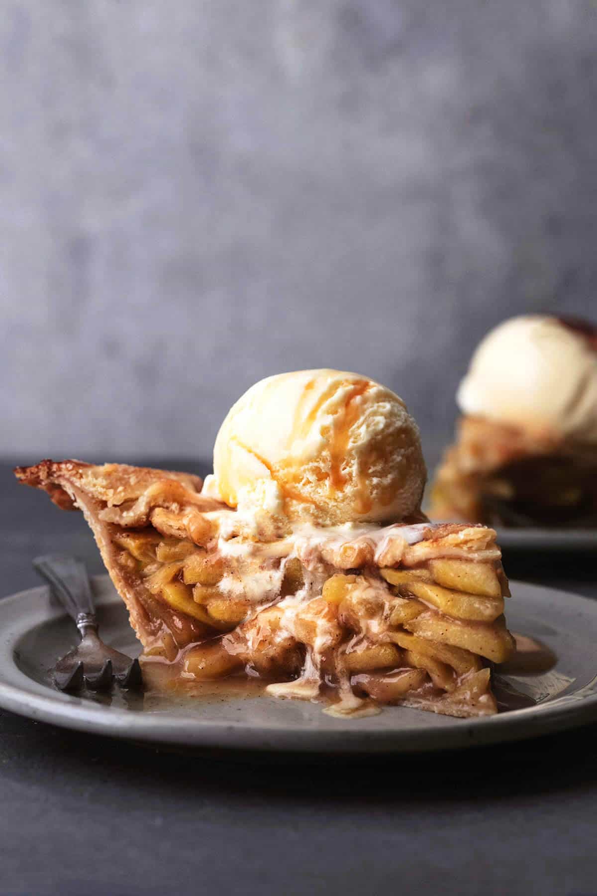 tabletop view of a slice of apple pie with an ice cream scoop on top on a plate with a fork.