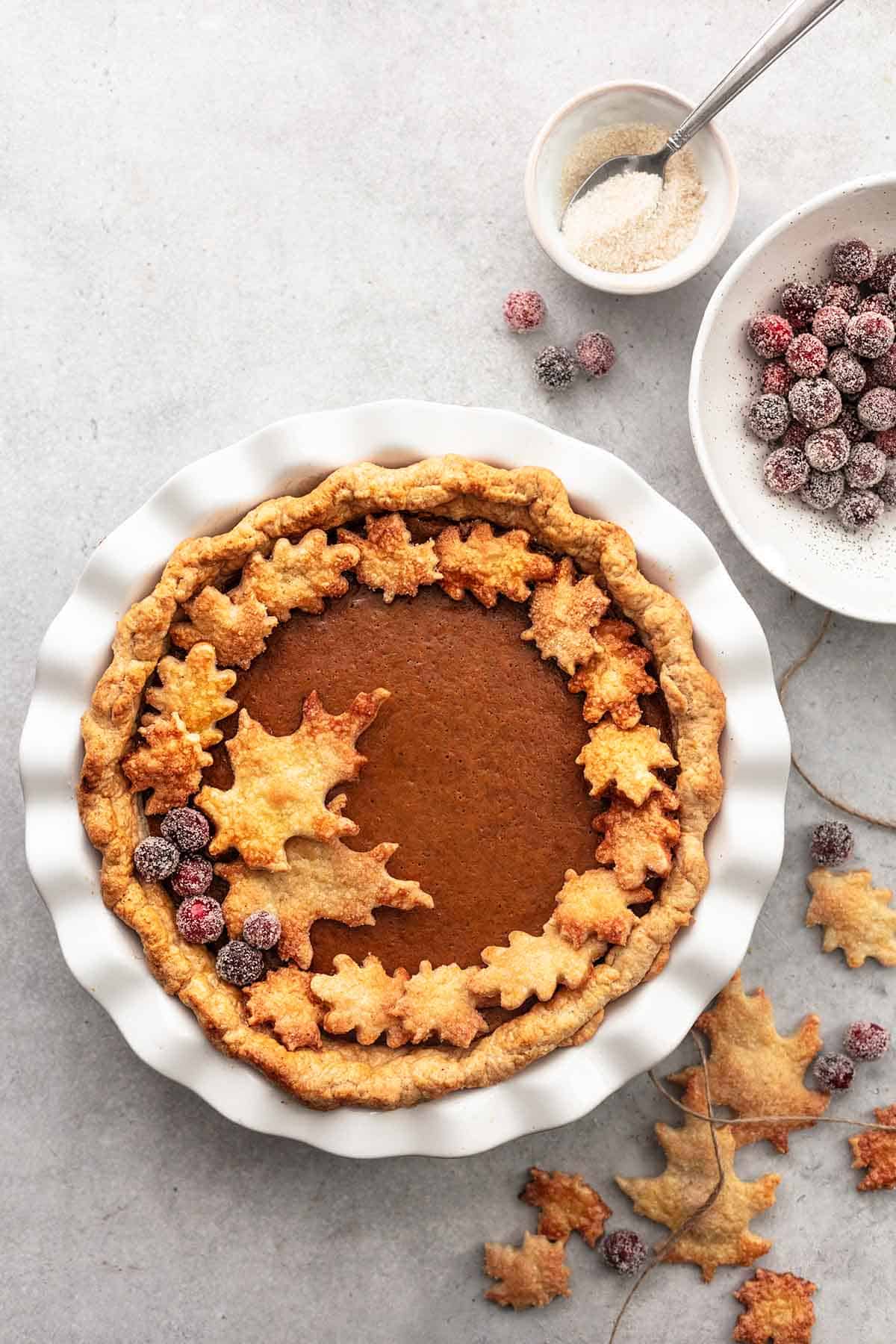 top view of pumpkin pie in a pie pan with sugar, cranberries in bowls and pie crust ornaments on the side.