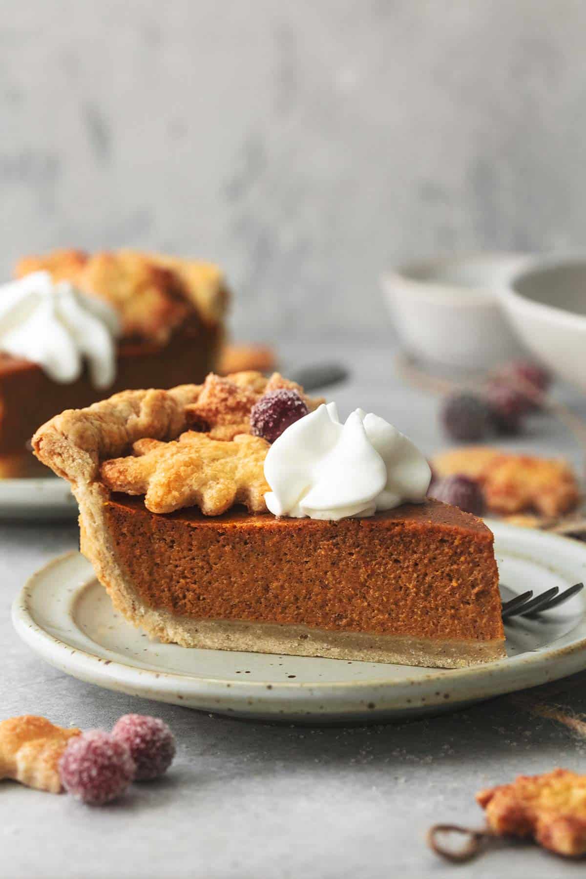 a slice of pumpkin pie topped with whipped cream and. sugared cranberries on a plate with a fork in front of another slice on a plate.