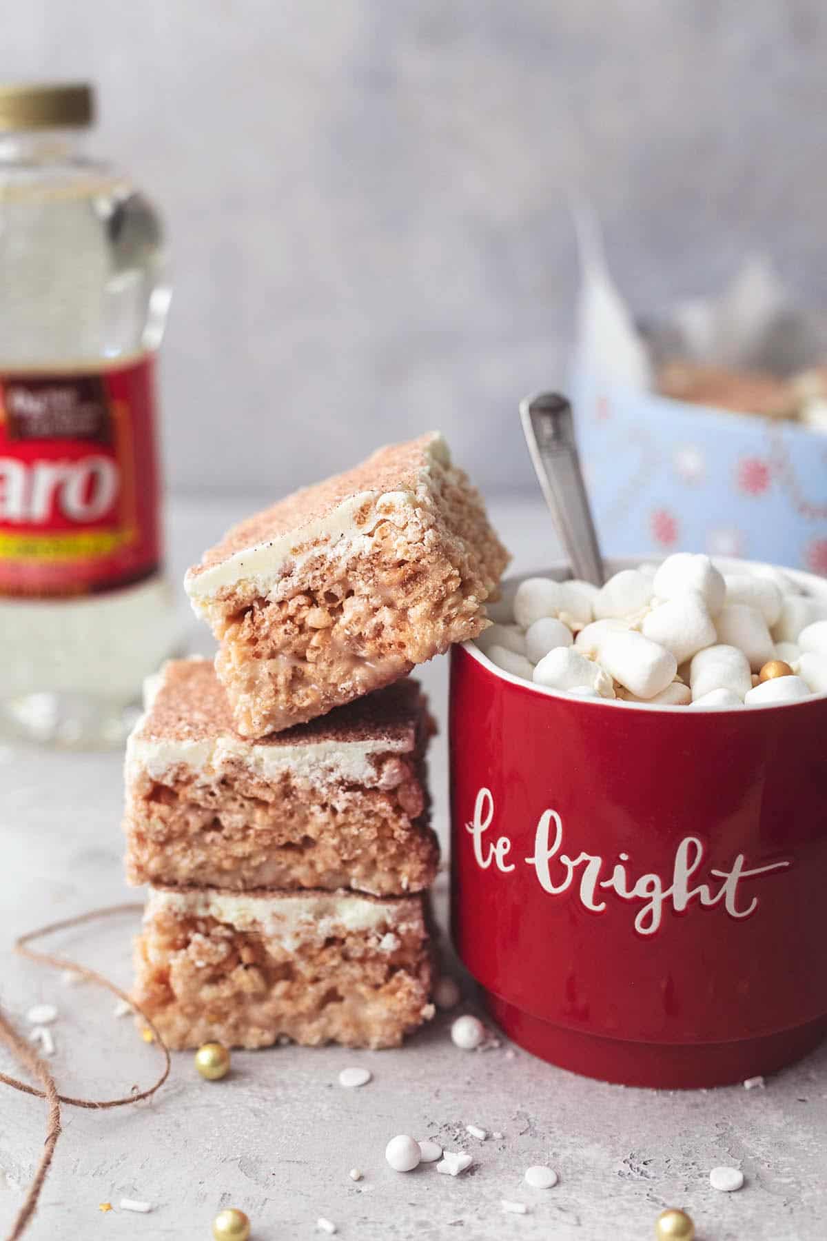 a stack of churro Krispie bars with the top bar leaning on a mug of hot chocolate with a bottle of Karo Corn Syrup and a holiday tin of more bars in the background.