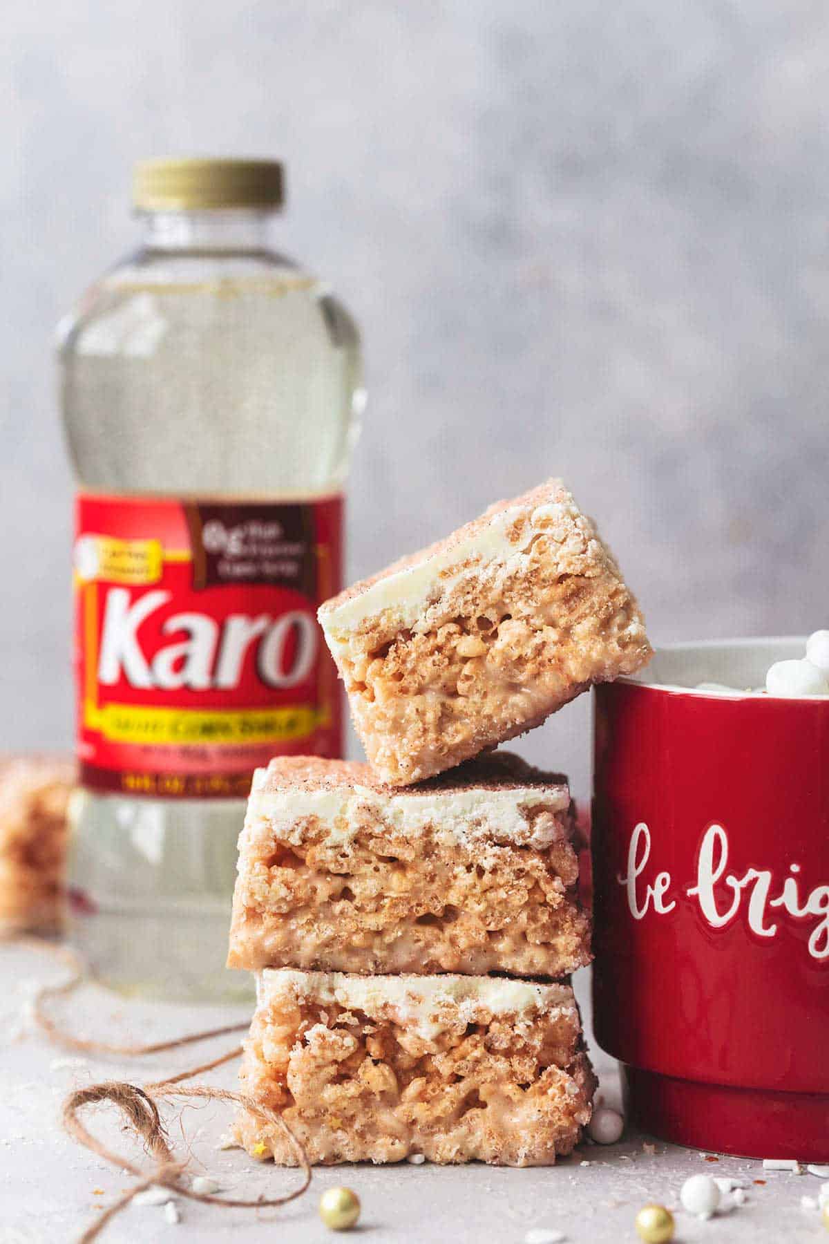 three stacked churro Krispie bars with white chocolate with the top bar leaning on a mug of hot chocolate with a bottle of Karo corn syrup in the background.