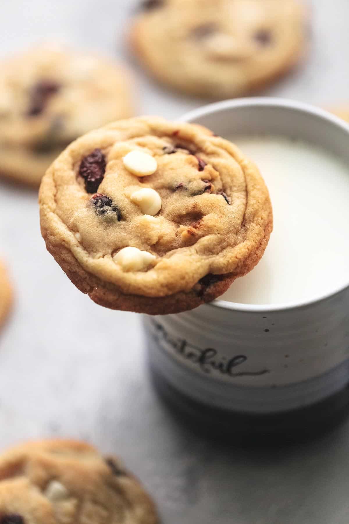 close up of a white chocolate cranberry cookie sitting on a mug of milk with more cookies around it.