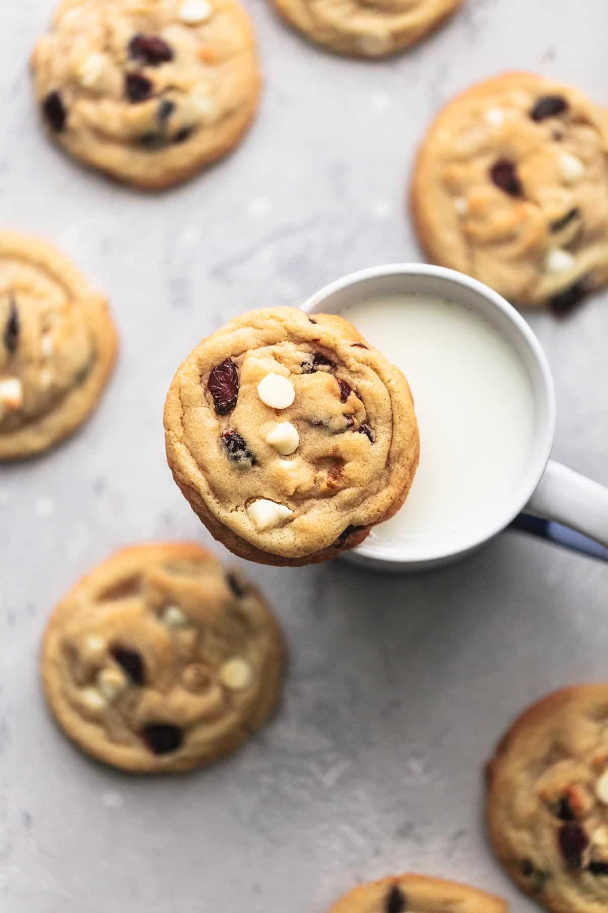 top view of a white chocolate cranberry cookie on top of a mug of milk with more cookies on the side.