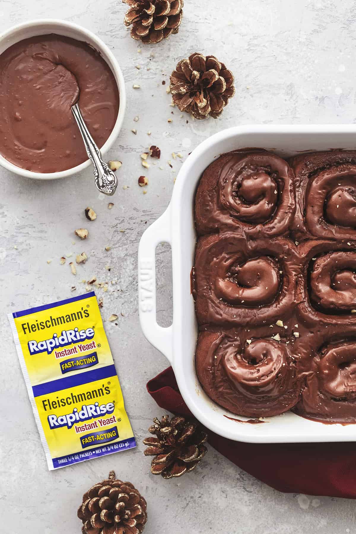 top view of chocolate cinnamon rolls with hazelnut icing in a baking pan with more icing in a bowl, yeast packets, and pinecones on the side.