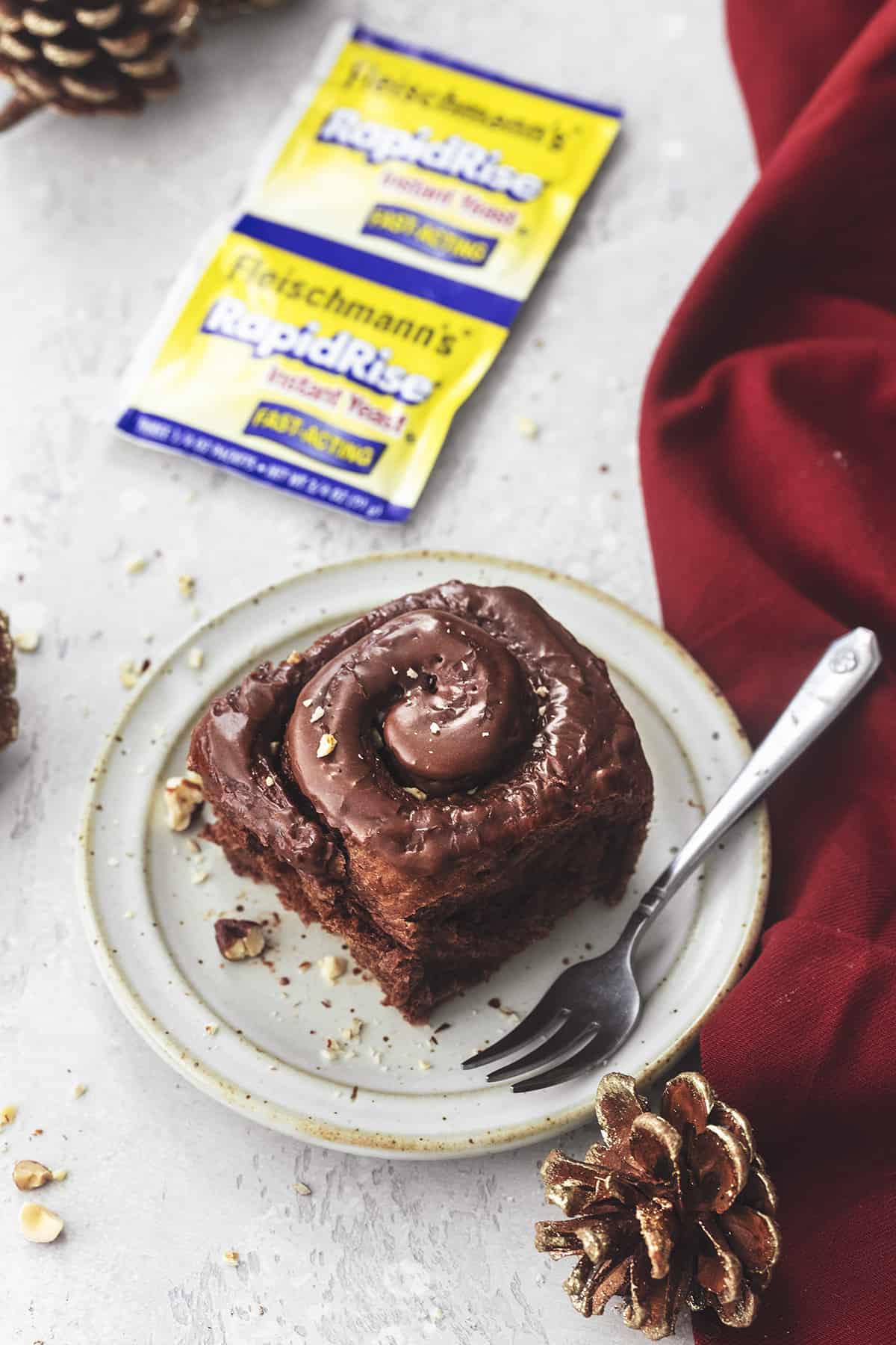 a chocolate cinnamon roll with hazelnut icing with a fork on a plate with pinecones and yeast packets on the side.