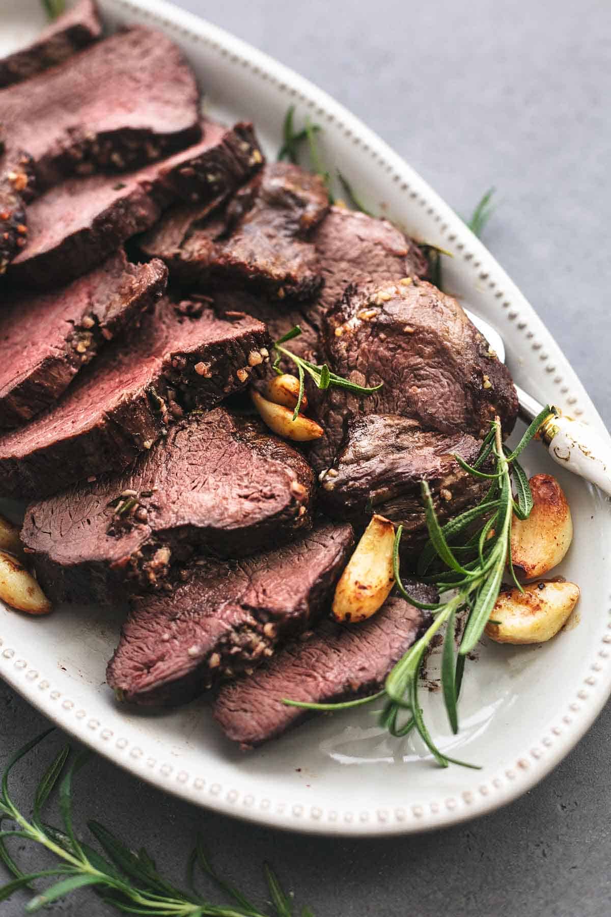 close up of sliced roasted garlic butter beef tenderloin on a platter with whole garlic cloves and rosemary.