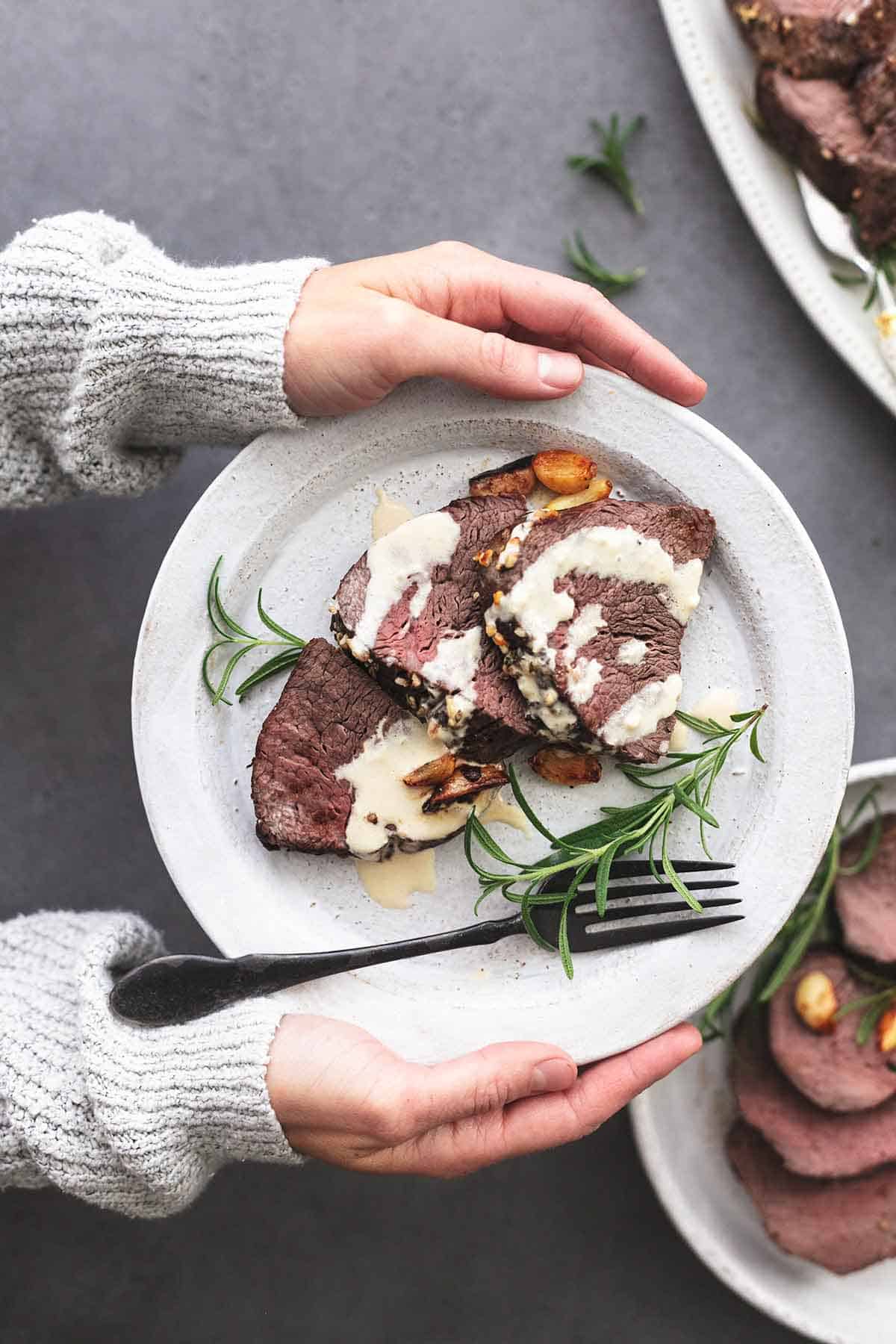 top view of hands holding a white plate with sliced roasted garlic butter beef tenderloin and fresh rosemary with a black fork.
