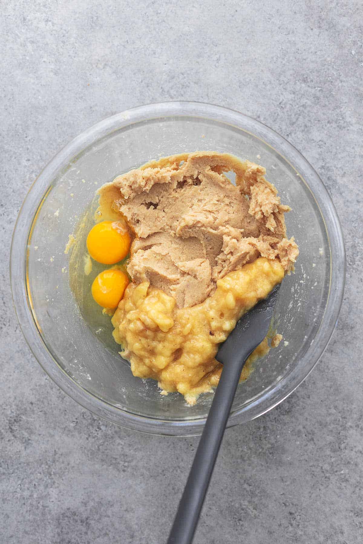 top view of mashed banana, creamy brown mixture, and egg yolks in a glass bowl with a spatula.