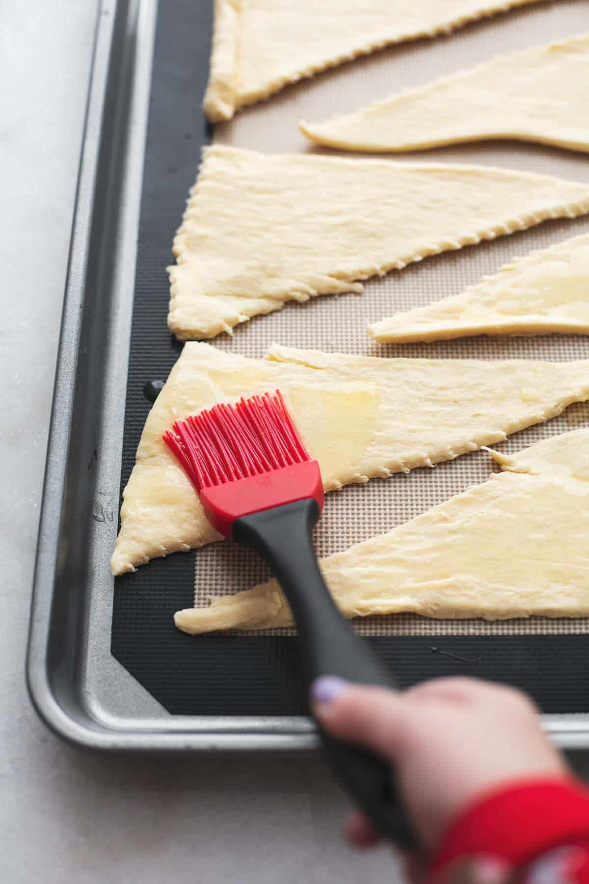 a child's hand brushing melted butter onto dough triangles on a baking sheet.