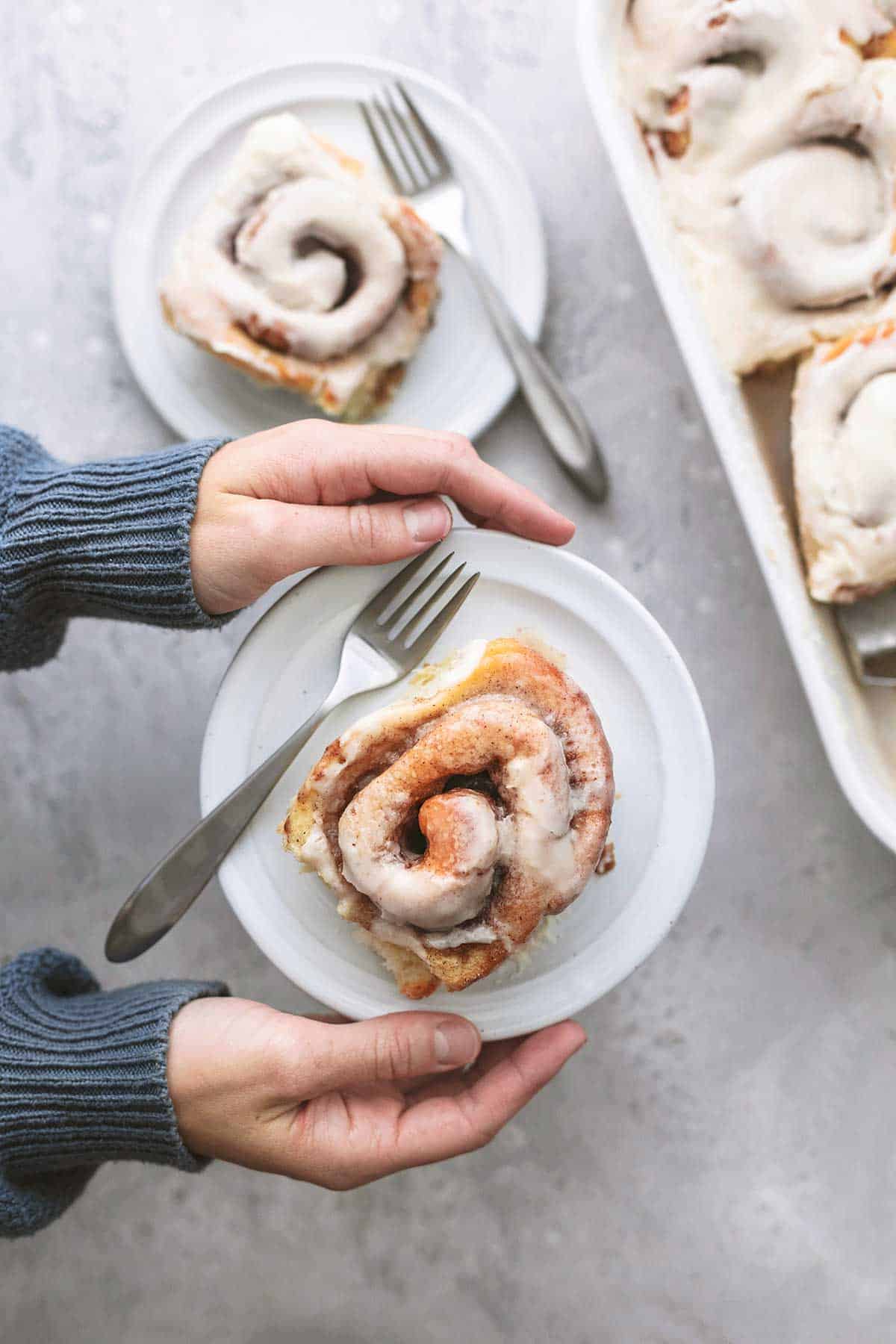 top view of hands holding a plate with a cinnamon roll with another plate and baking pan of rolls on the side.
