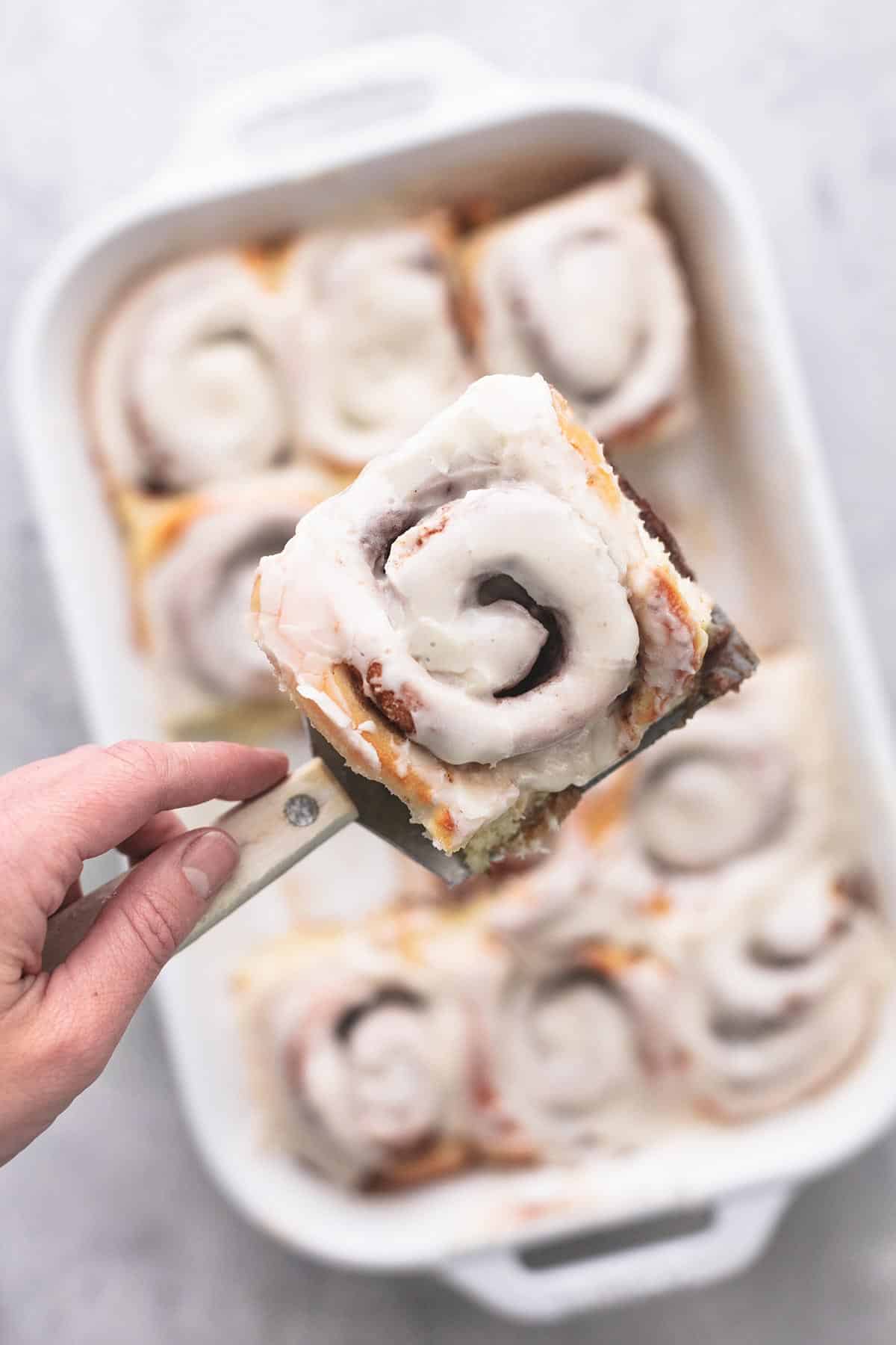 top view of a hand holding a spatula with a cinnamon roll on top above a pan of more rolls.